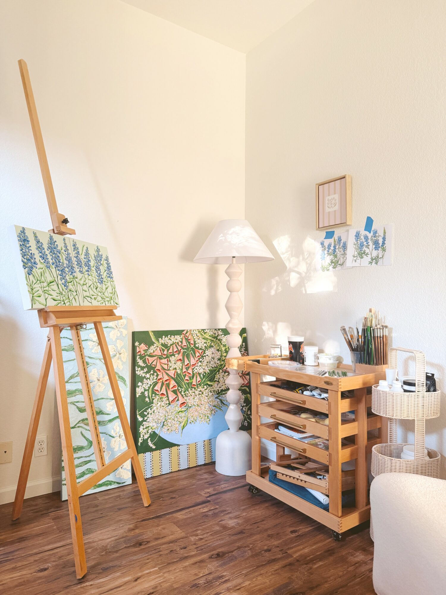 Corner of a room with an easel, paintings, a lamp, and a wooden storage unit, with a wooden floor and white walls.