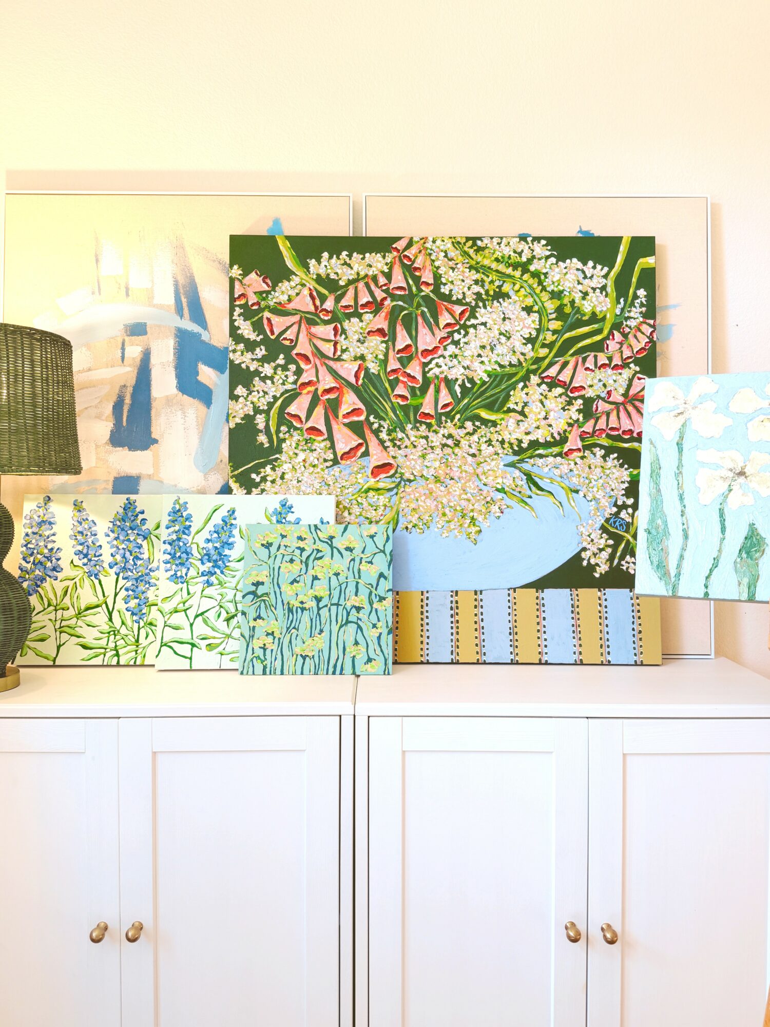 Multiple colorful paintings of flowers and plants displayed on a white cabinet, with a beige wall in the background.