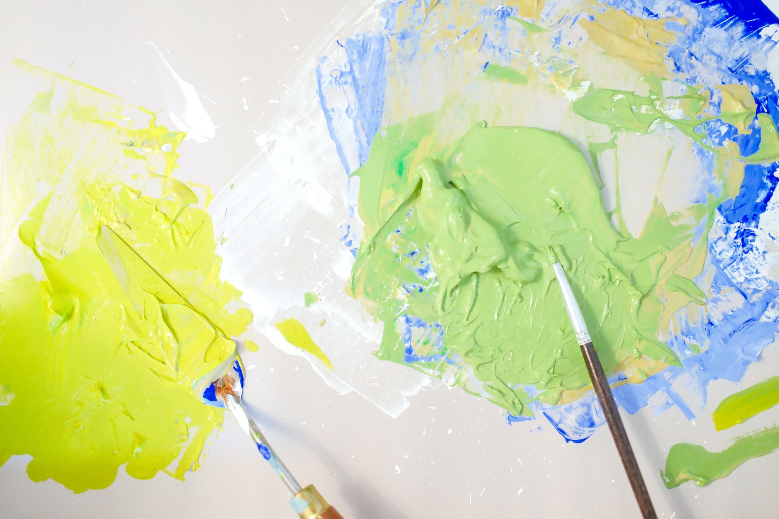 Two paint palettes with yellow and green paint, brushes resting on them, on a white surface.