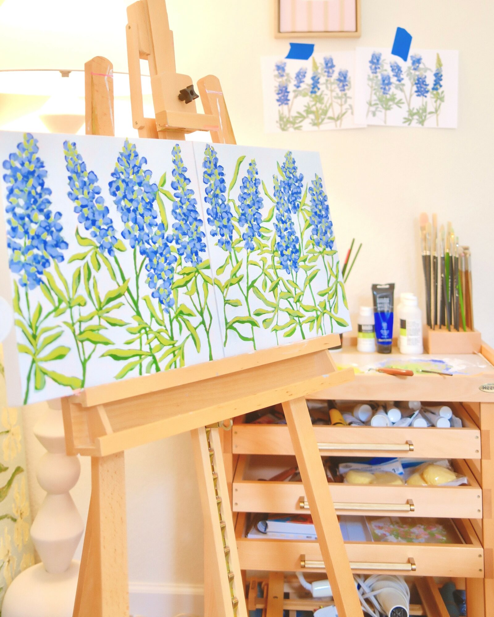 Three paintings of blue flowers on an easel, with art supplies and sketches nearby, in a bright art studio.