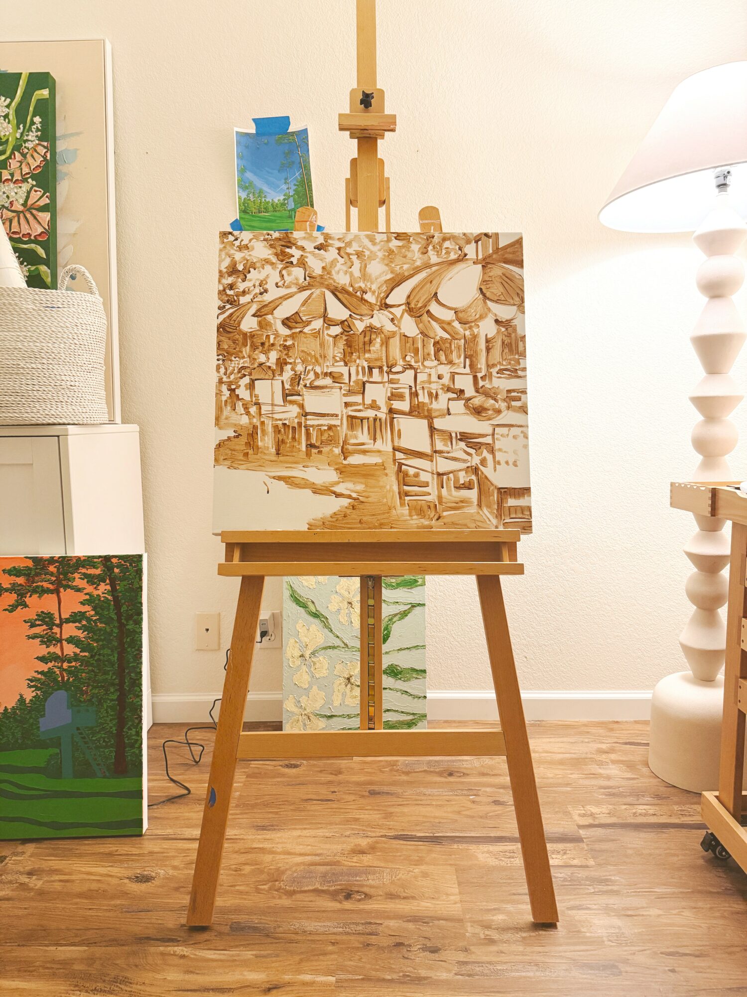 Easel with a painting of a cityscape, surrounded by other artworks and a lamp in a room.