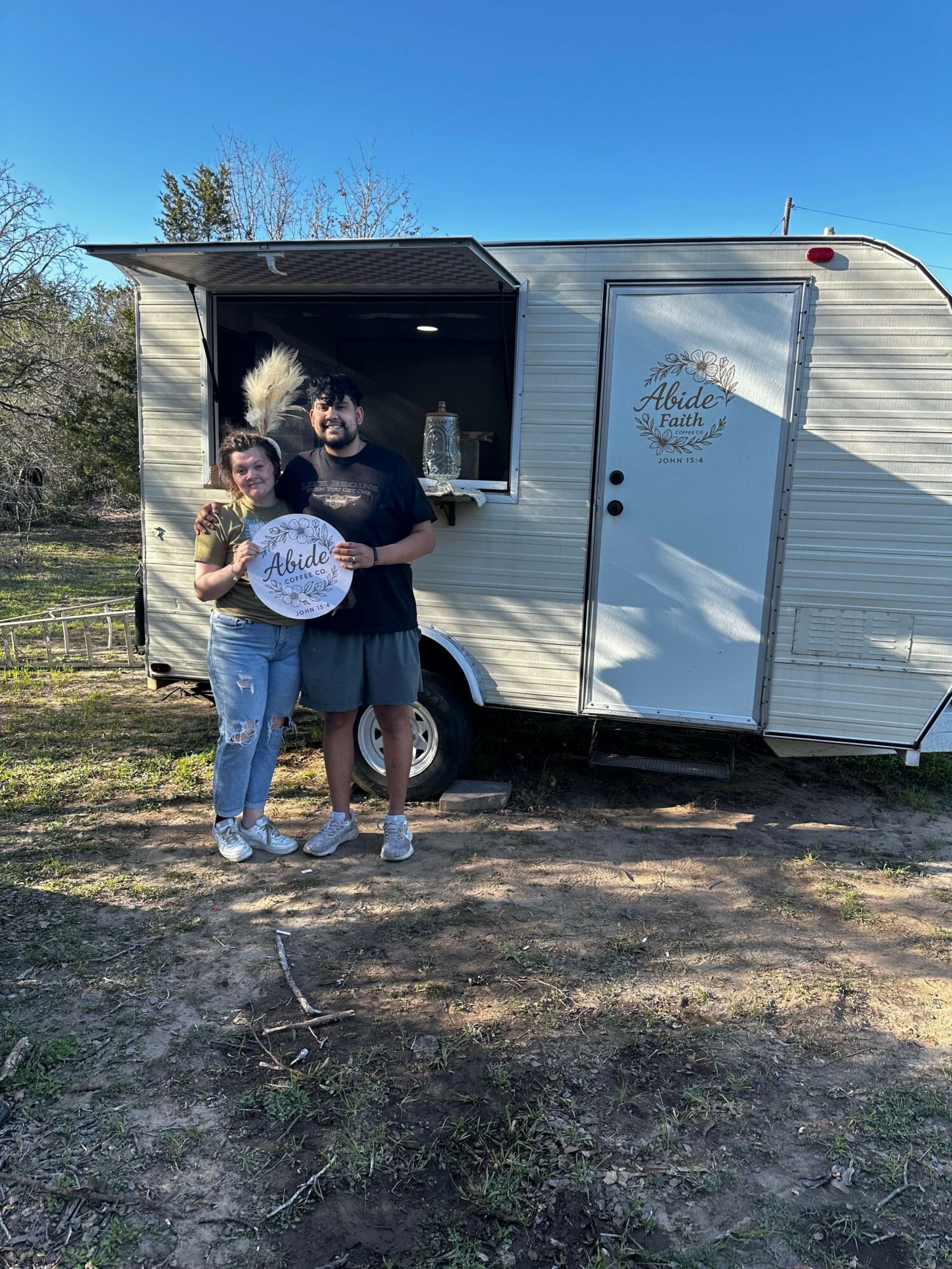 Person standing outside a small white trailer with an open door, holding a child and a dog, on a dirt ground.