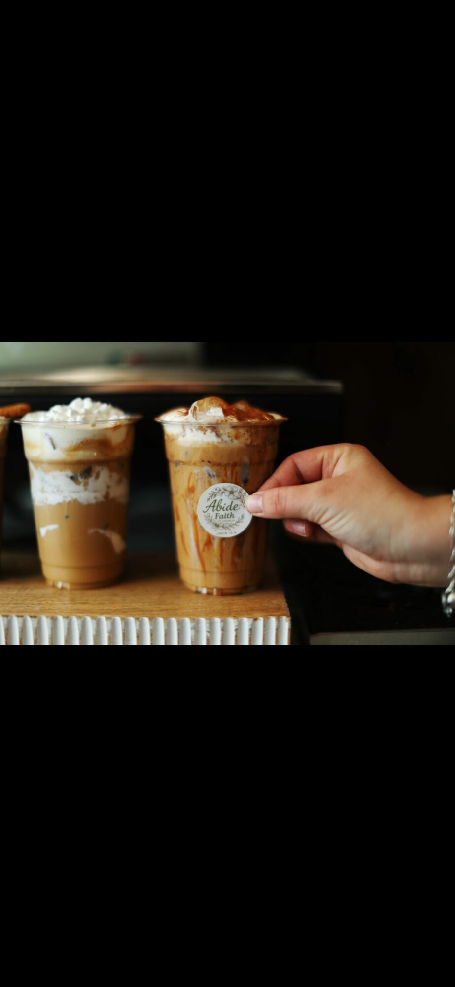 Person holding a small cup of coffee with whipped cream, next to a larger coffee drink with whipped cream on a wooden surface.