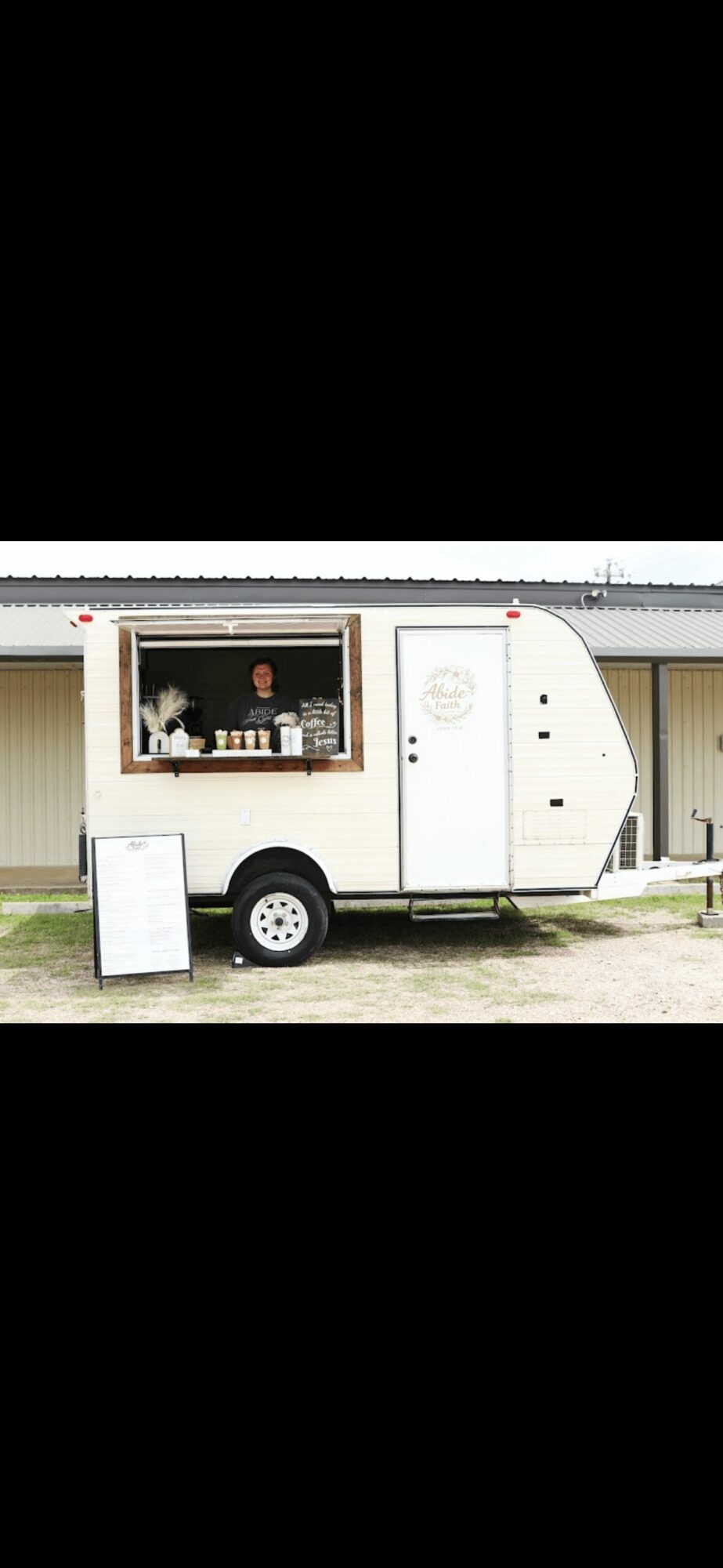 Small food truck with a window serving food, parked outside a building with a yellow wall and black roof.