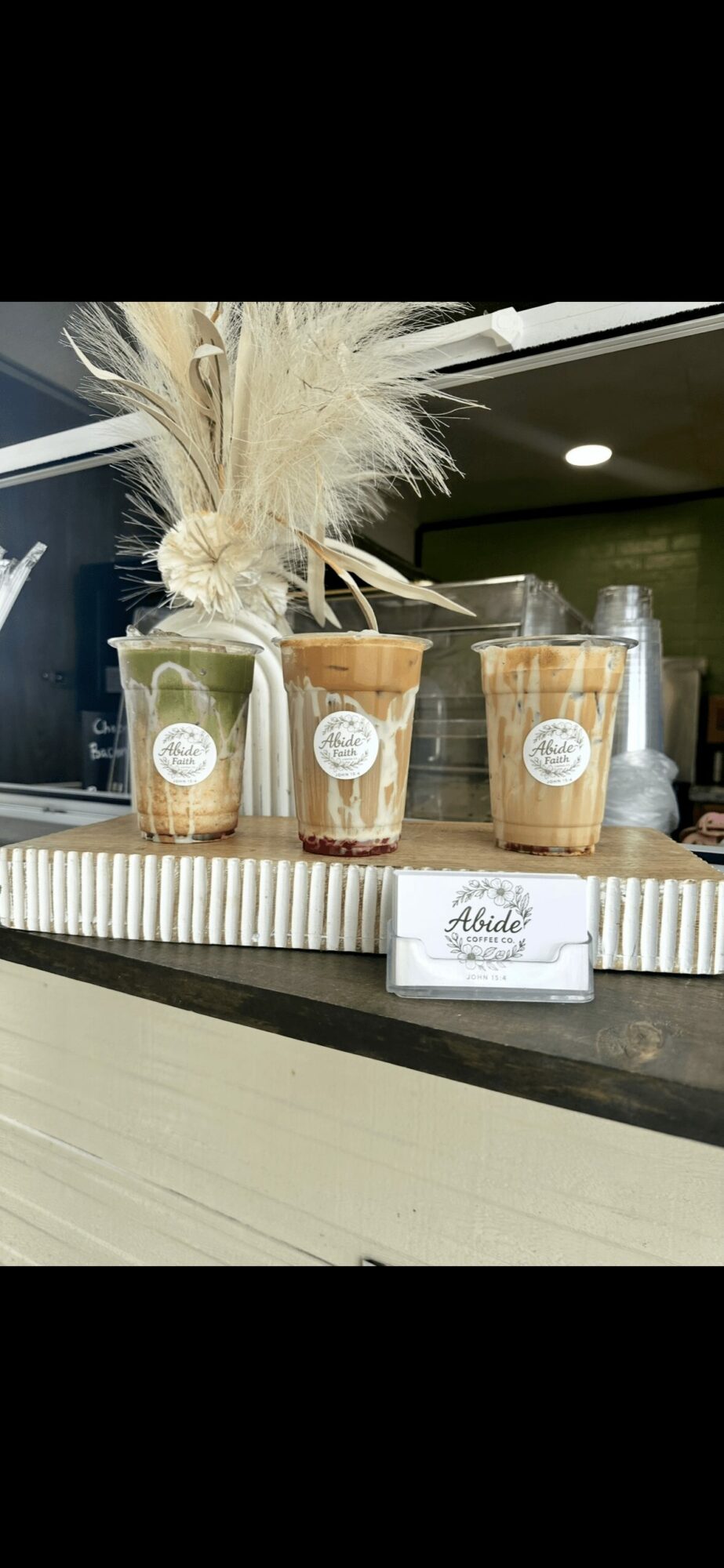 Three clear cups with labels and a plant with tall, feathery beige plumes on a counter, with a small sign in front.