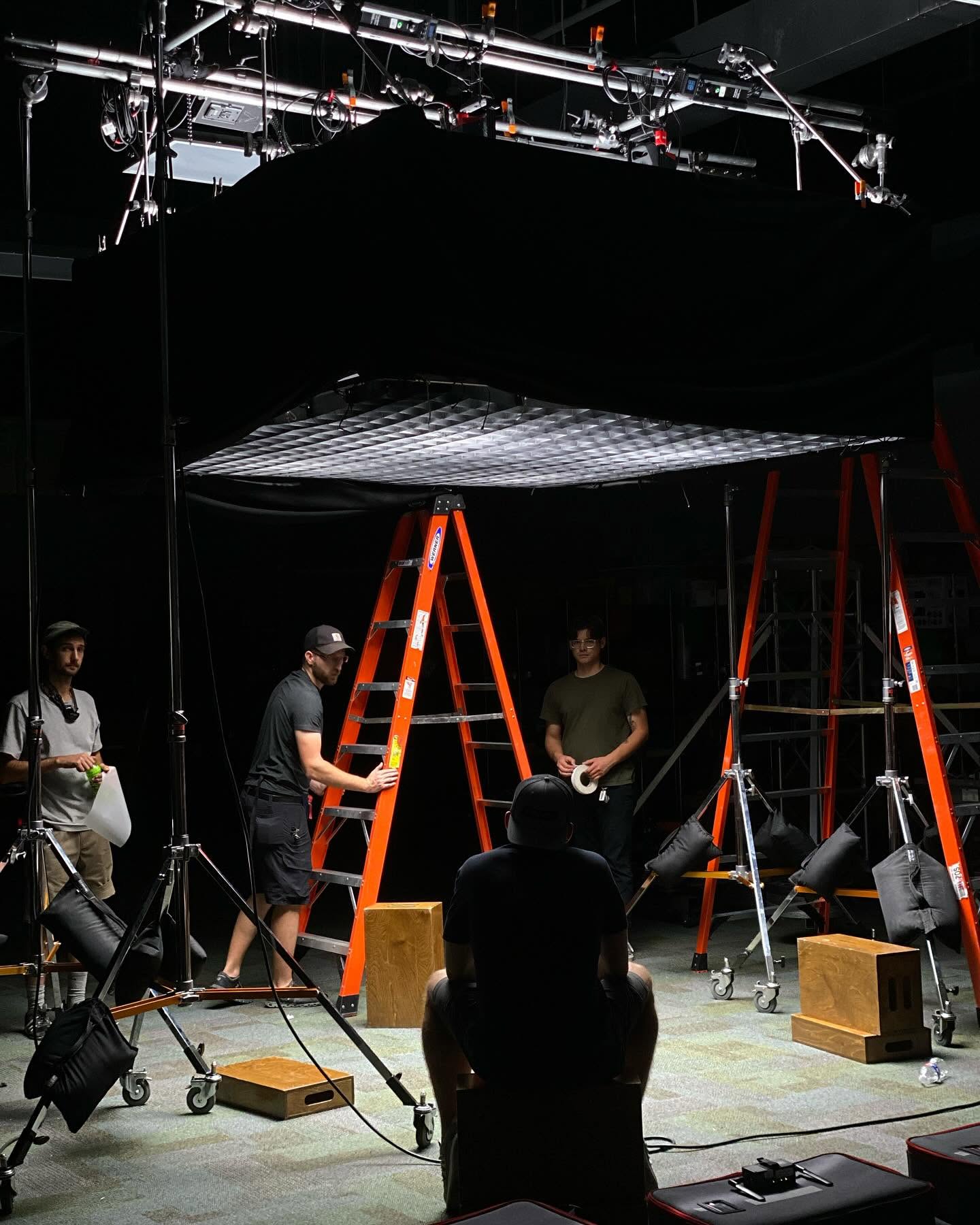 People working on a stage setup with ladders, lighting, and equipment in a dark room.