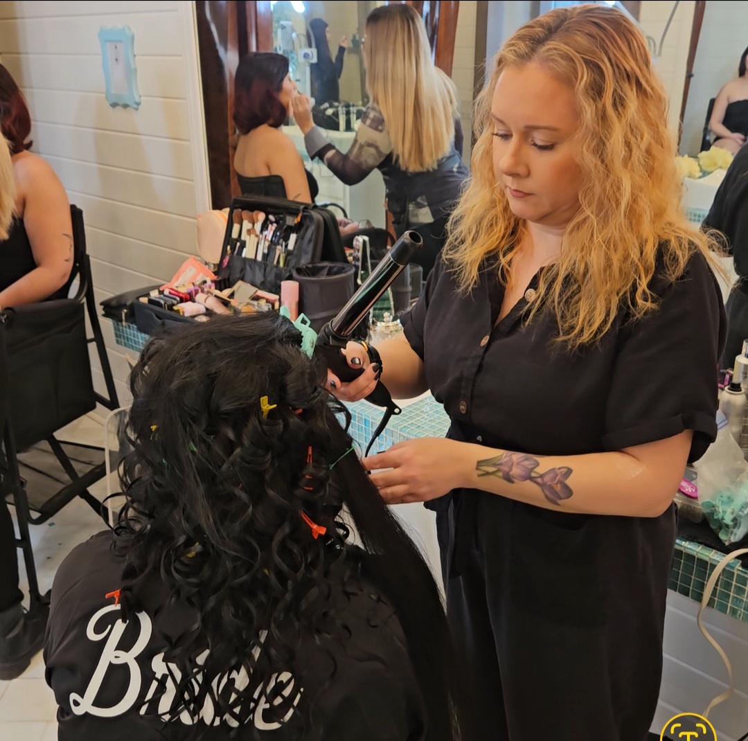 A hairstylist with curly blonde hair and a tattoo on her arm styles a client's long, curly hair in a salon.
