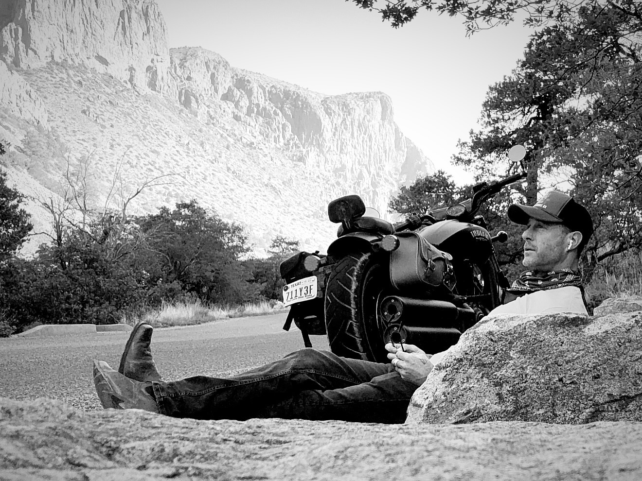 Person lying on the ground next to a motorcycle, with a mountain and trees in the background.