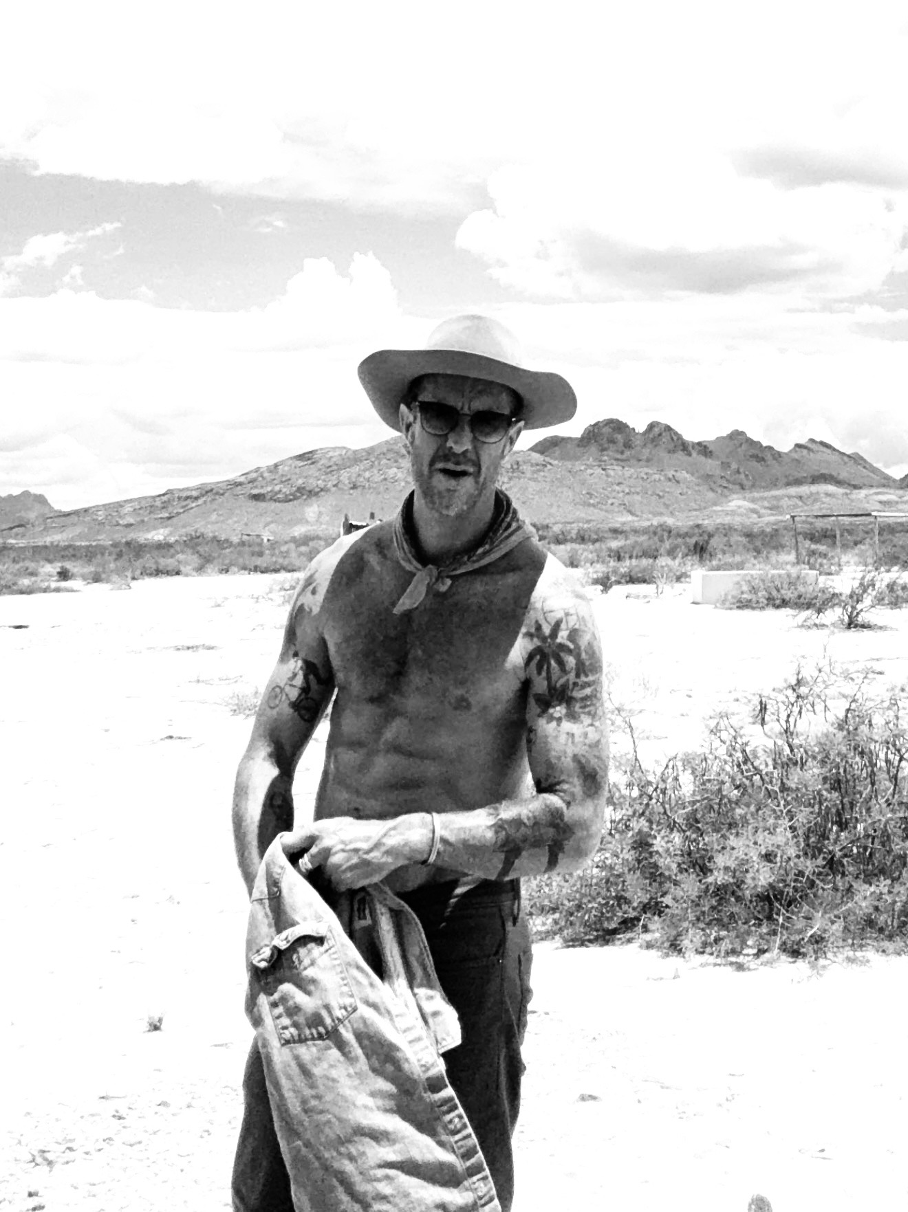 Man standing outdoors in a desert landscape, wearing a hat, sunglasses, and a bandana, holding a jacket, with mountains in background.