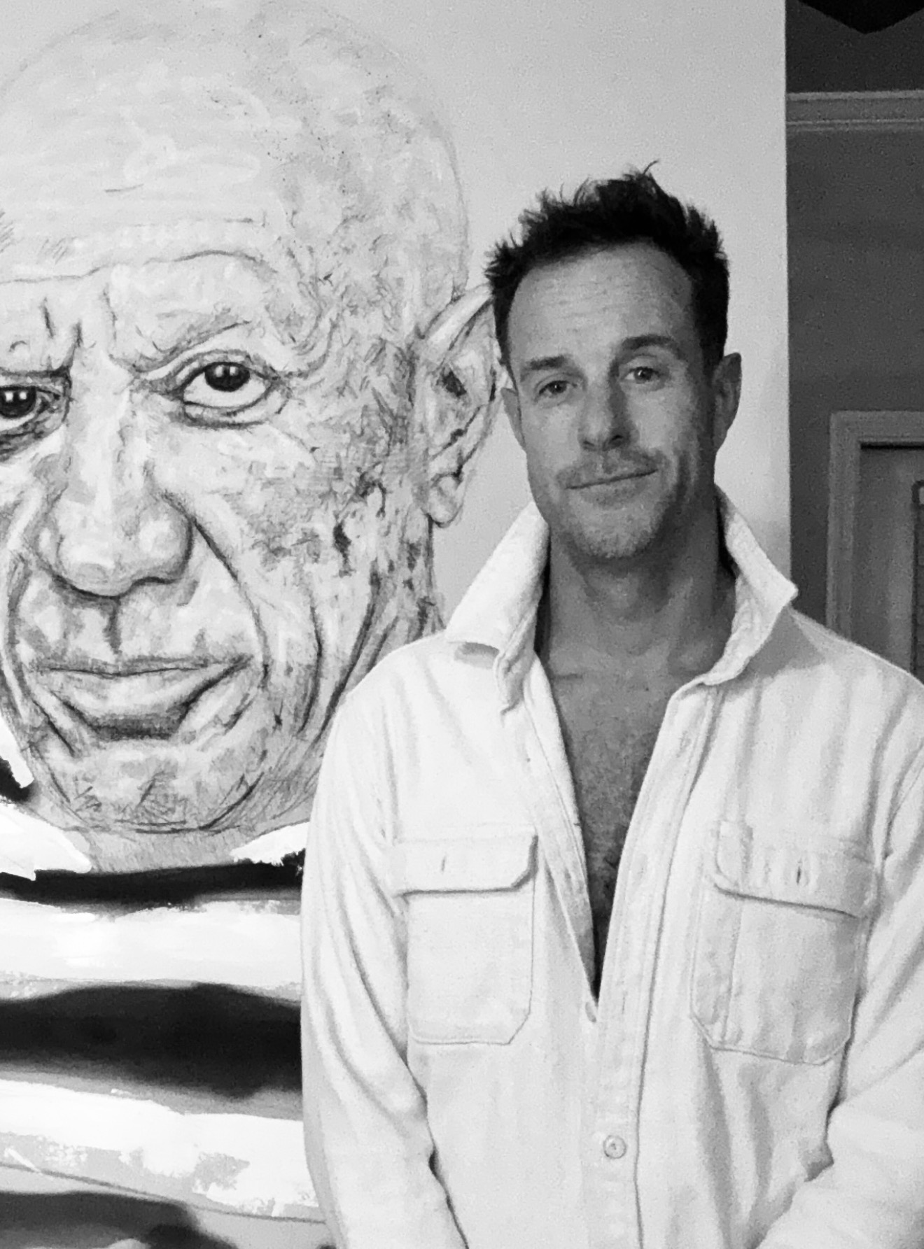 Man with short dark hair and a slight smile, wearing a white shirt, standing next to a large portrait of an elderly person's face.