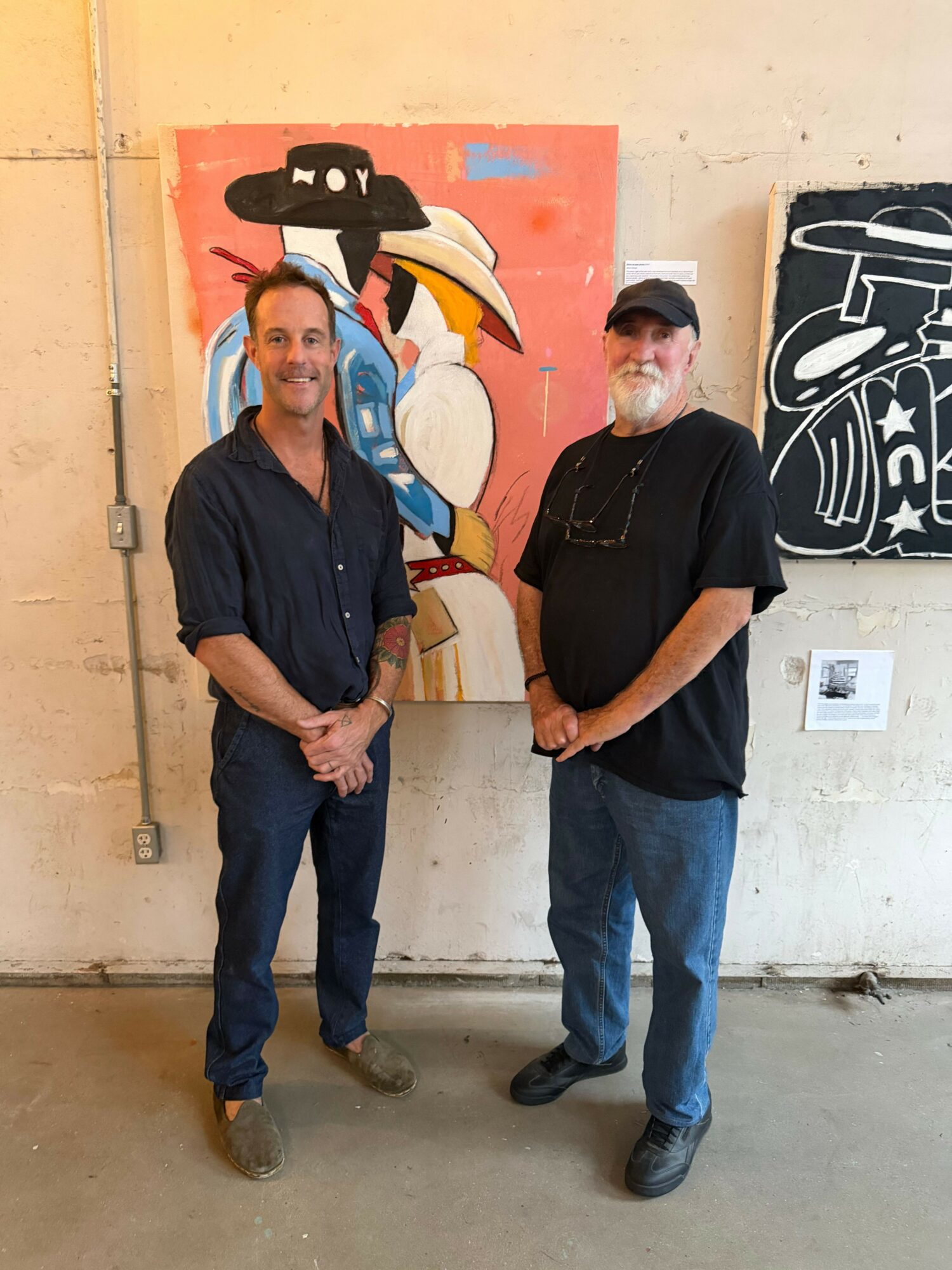 Two men standing in front of a colorful abstract painting on a wall, one wearing a black shirt and jeans, the other in a dark jacket and pants.