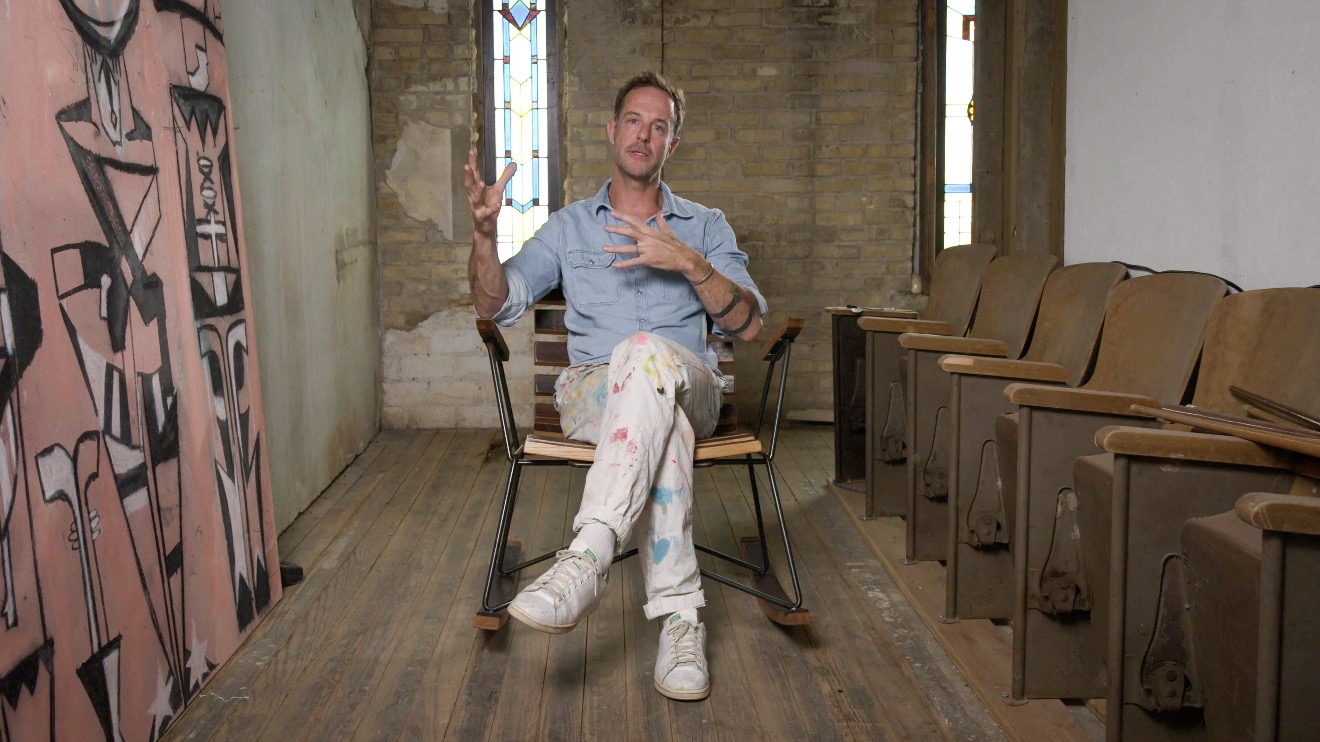 Man sitting on a chair in a room with wooden floors and chairs, gesturing with one hand, smiling, with stained glass window behind.