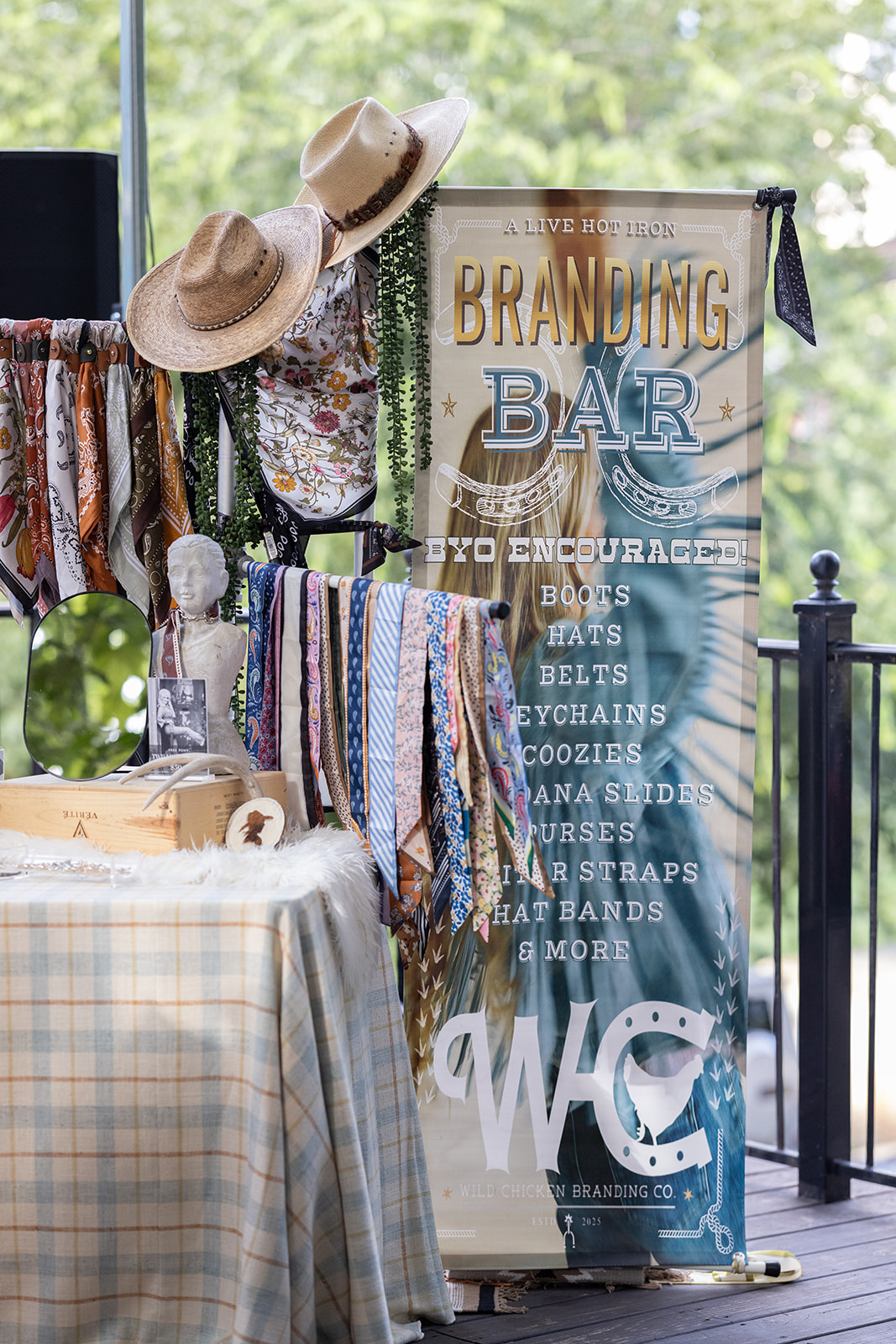 Outdoor market stall with hats, scarves, and a sign advertising a bar and food items, set against a green, leafy background.