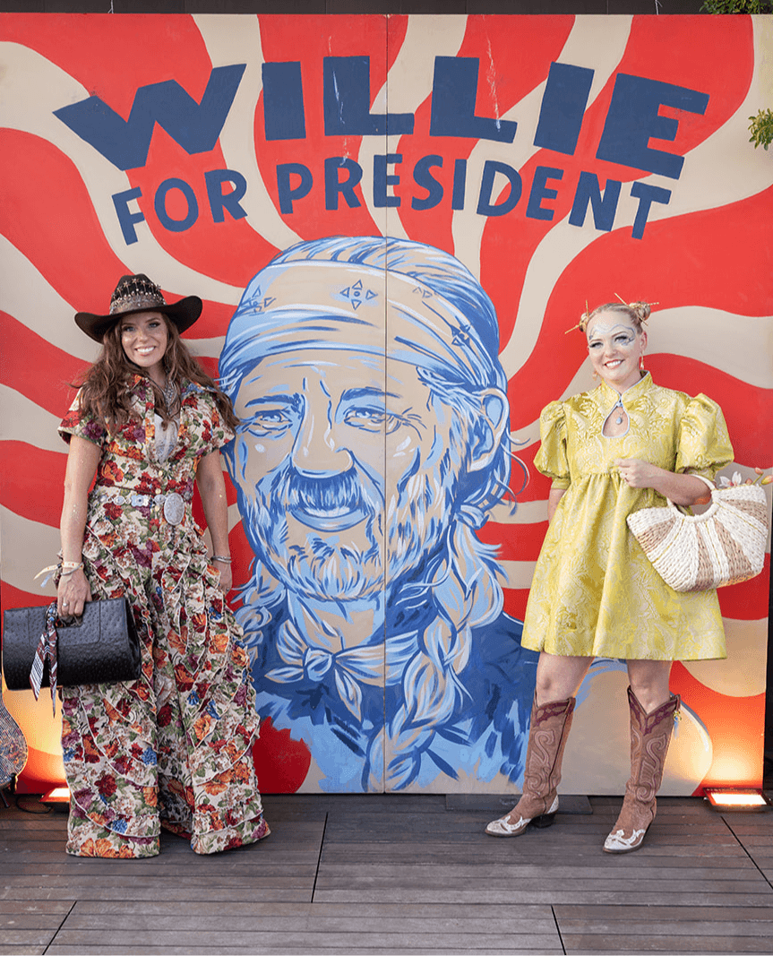 Two women stand in front of a colorful mural with a large portrait of Willie and the text 'Willie for President'.