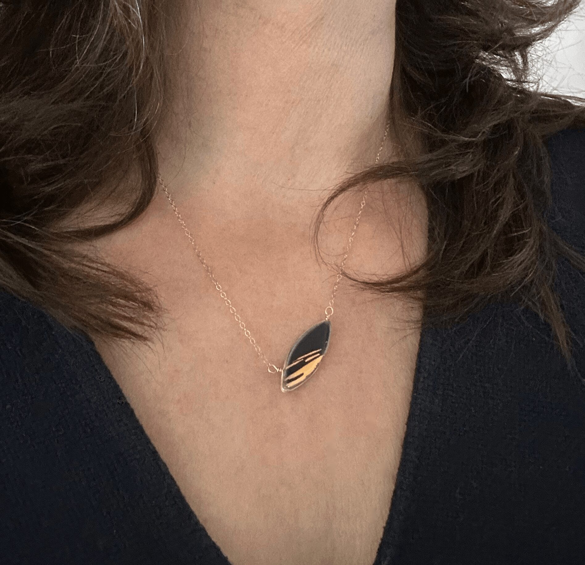 Person wearing a gold and black leaf-shaped pendant necklace on a chain, with dark hair and a black top.