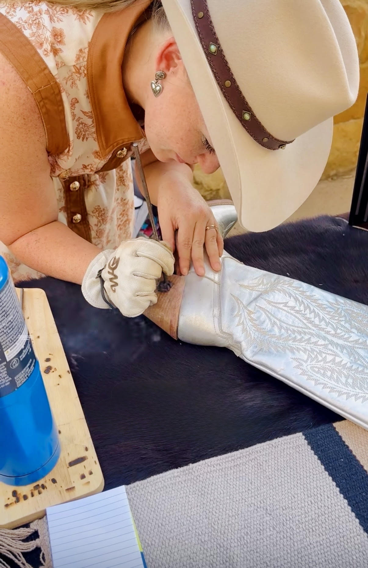 Person wearing a cowboy hat and gloves is working on a piece of leather or fabric, with a blue water bottle nearby.
