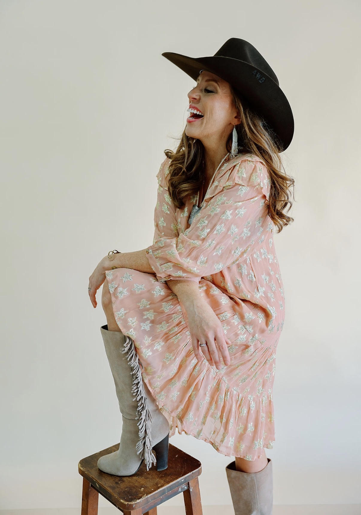 Woman wearing a large black hat and pink patterned dress, sitting on a stool with one foot on a boot, smiling.