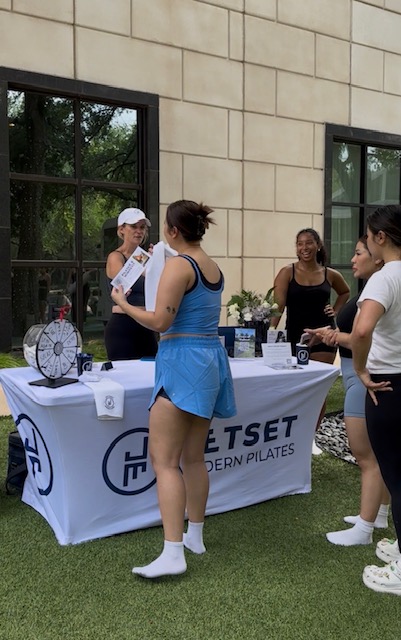 Group of people at an outdoor event with a table and promotional materials, in front of a building with large windows.