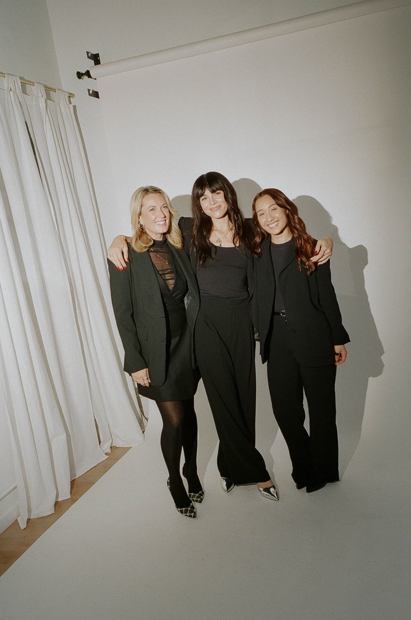 Three women in black outfits standing together, smiling, with arms around each other, in a white room with curtains.