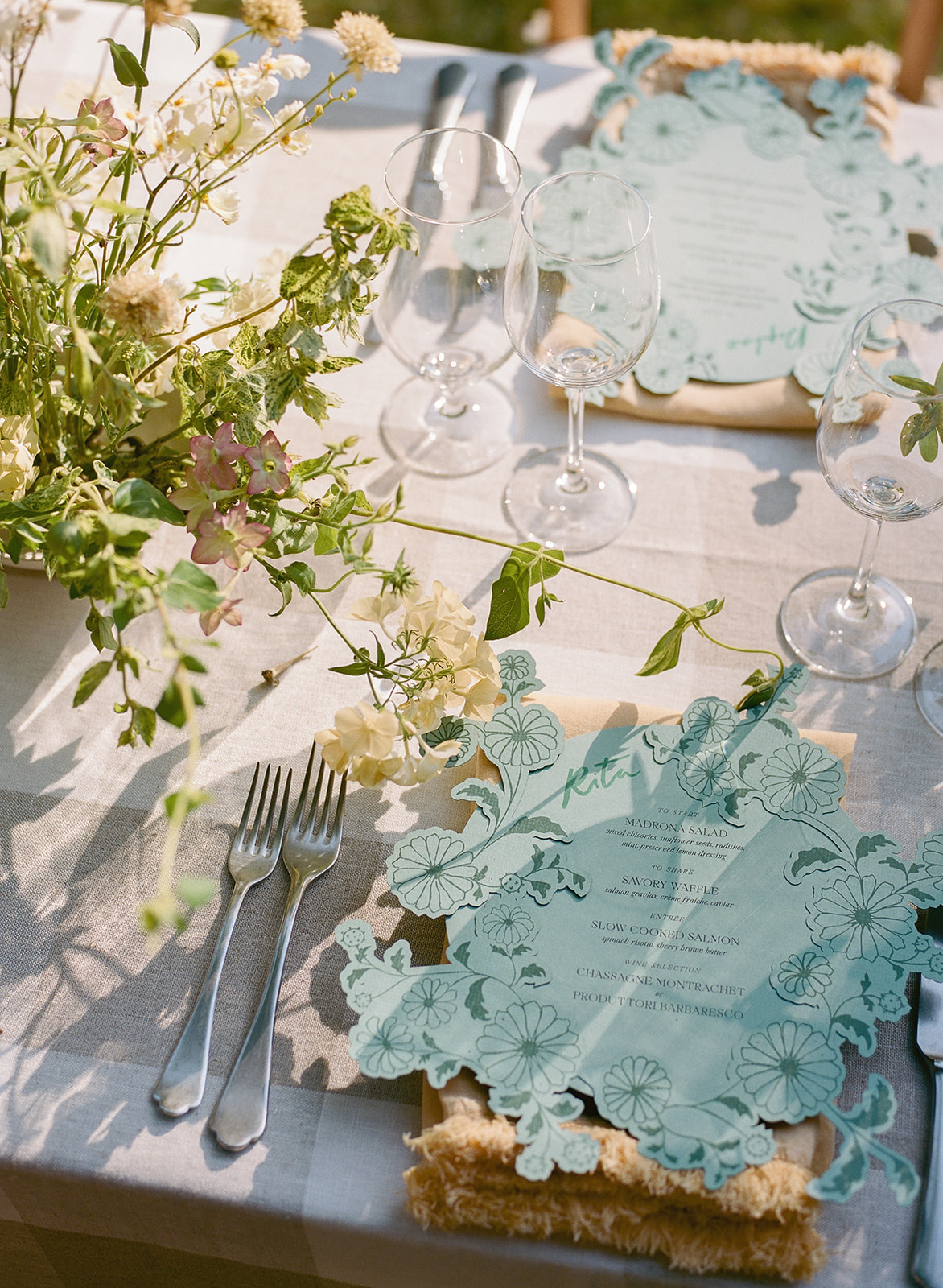 Table setting with floral centerpiece, wine glasses, and decorative blue placemats, outdoors with natural light.