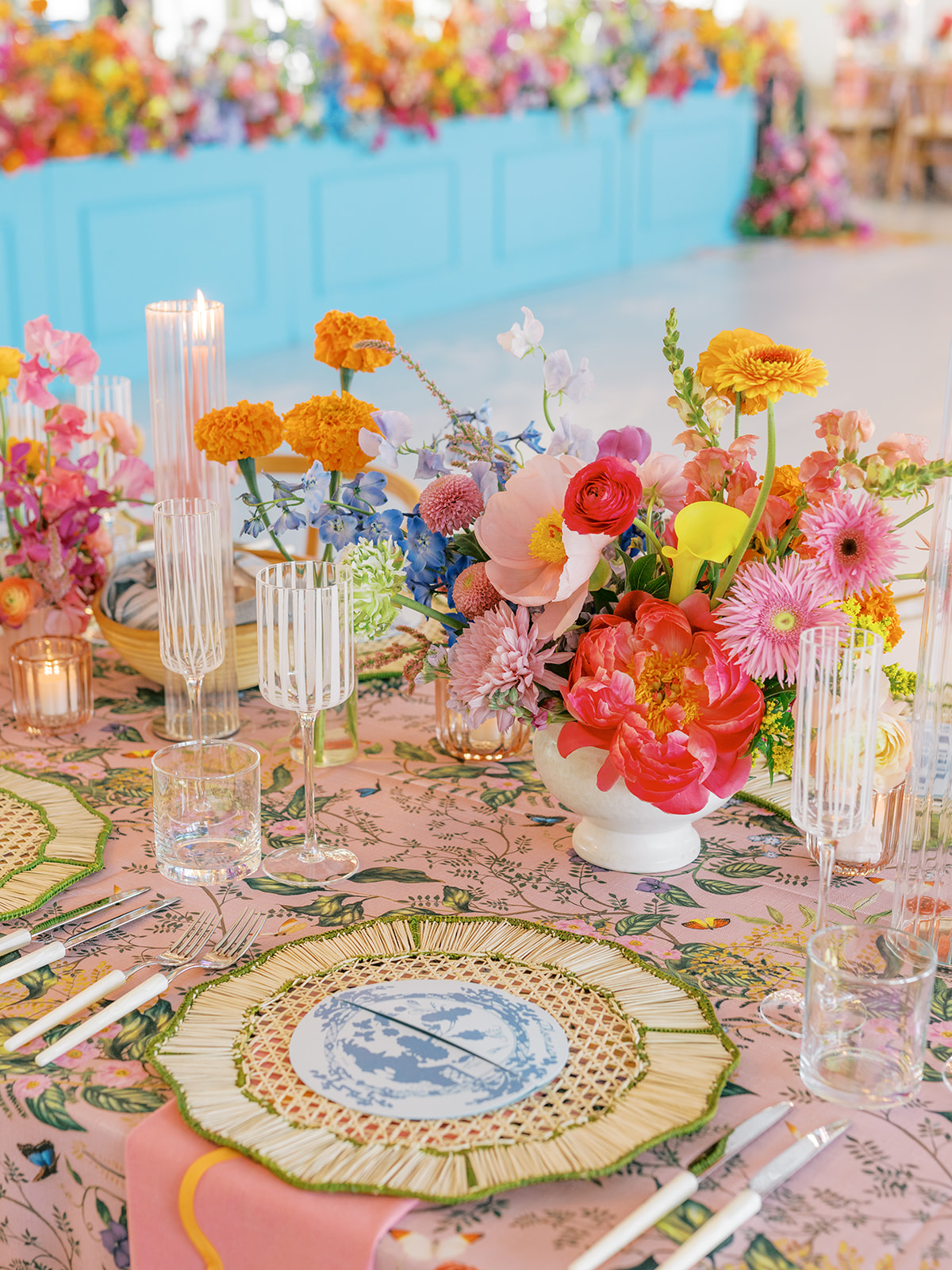Table decorated with colorful flowers, candles, and glassware, set for a celebration or event, with a pink tablecloth and vibrant background.