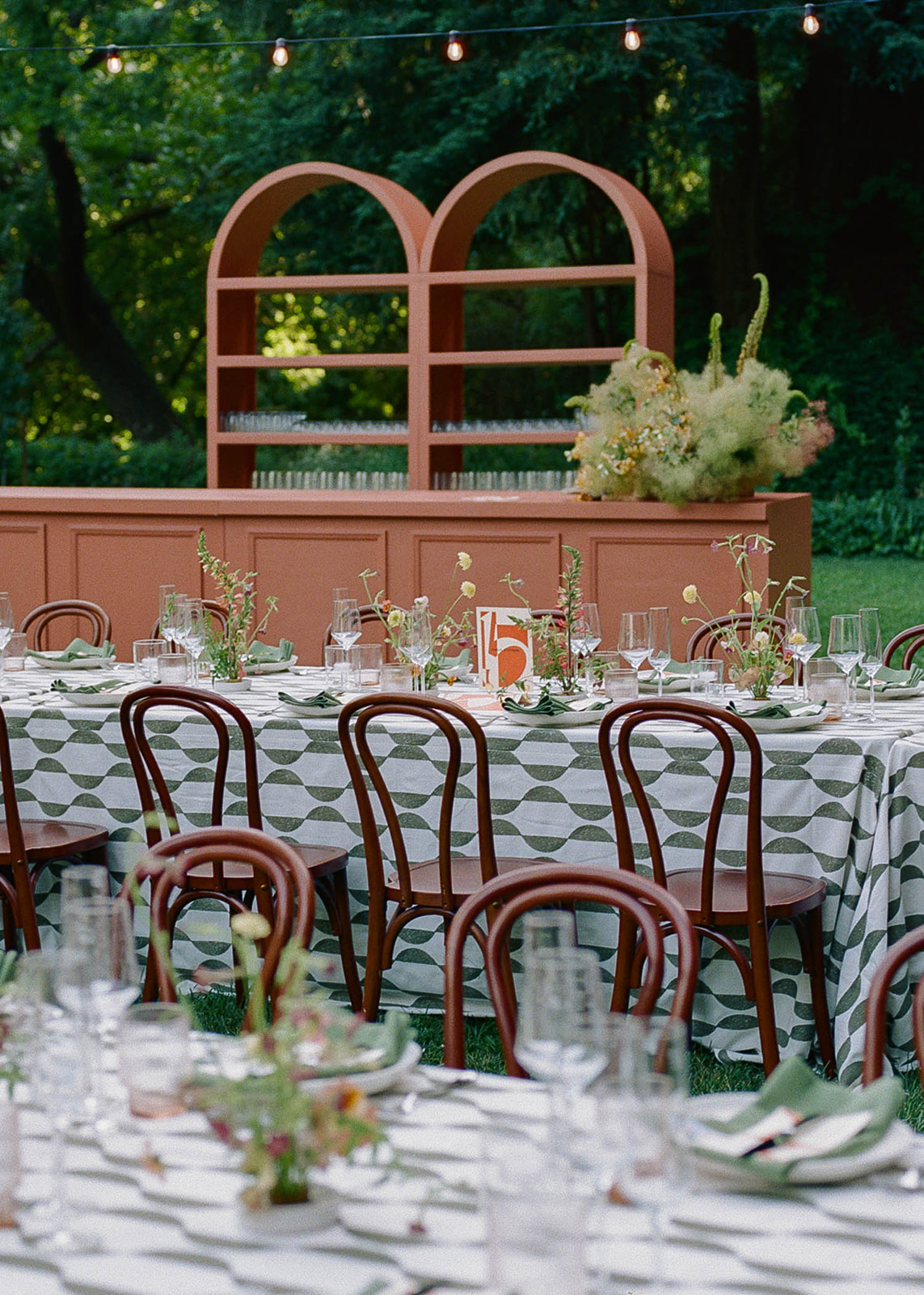 Outdoor table set with chairs, tablecloth, glasses, and floral centerpieces, with a pink shelf and greenery in background.