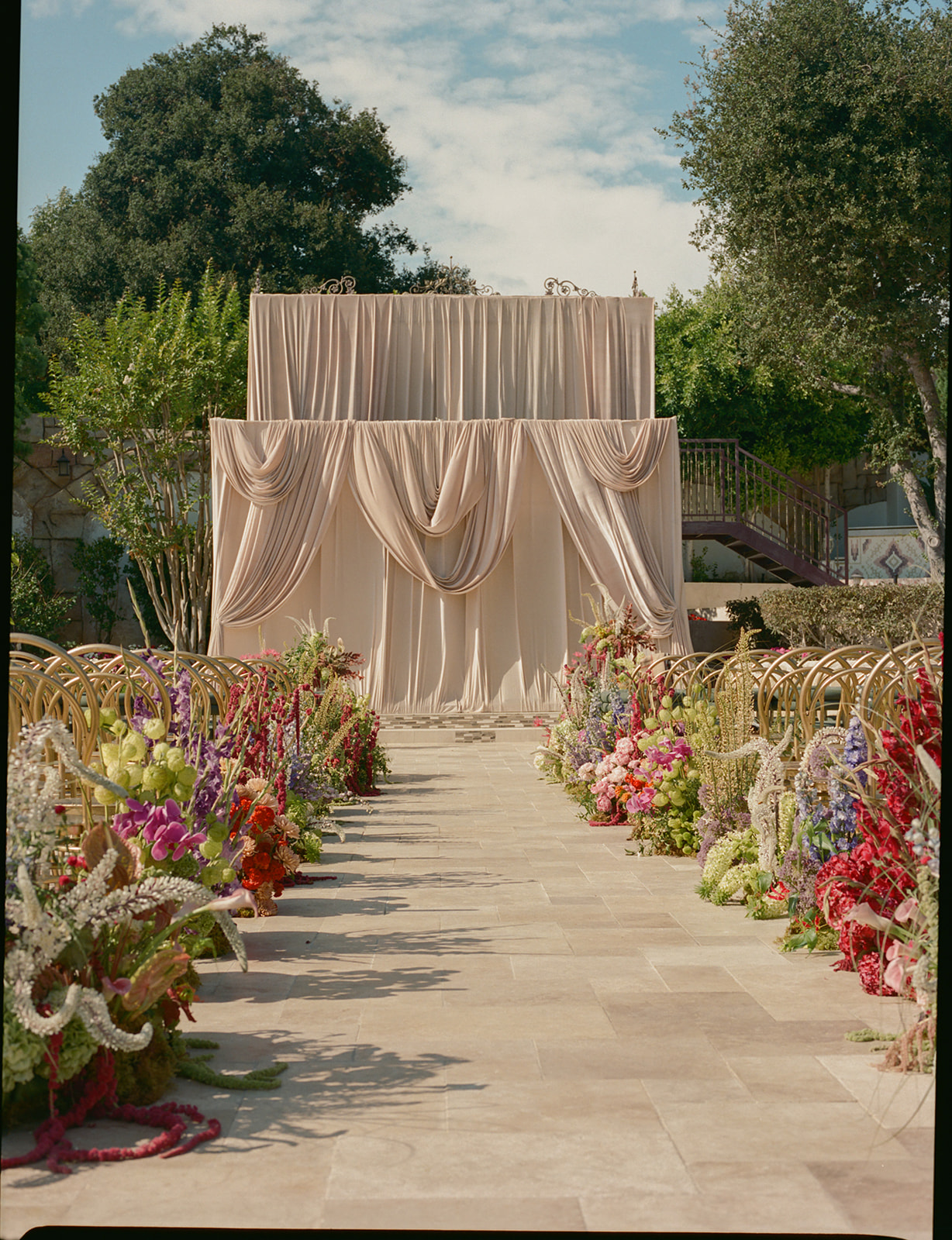 Outdoor wedding aisle with floral arrangements and a draped backdrop, trees and sky in background.