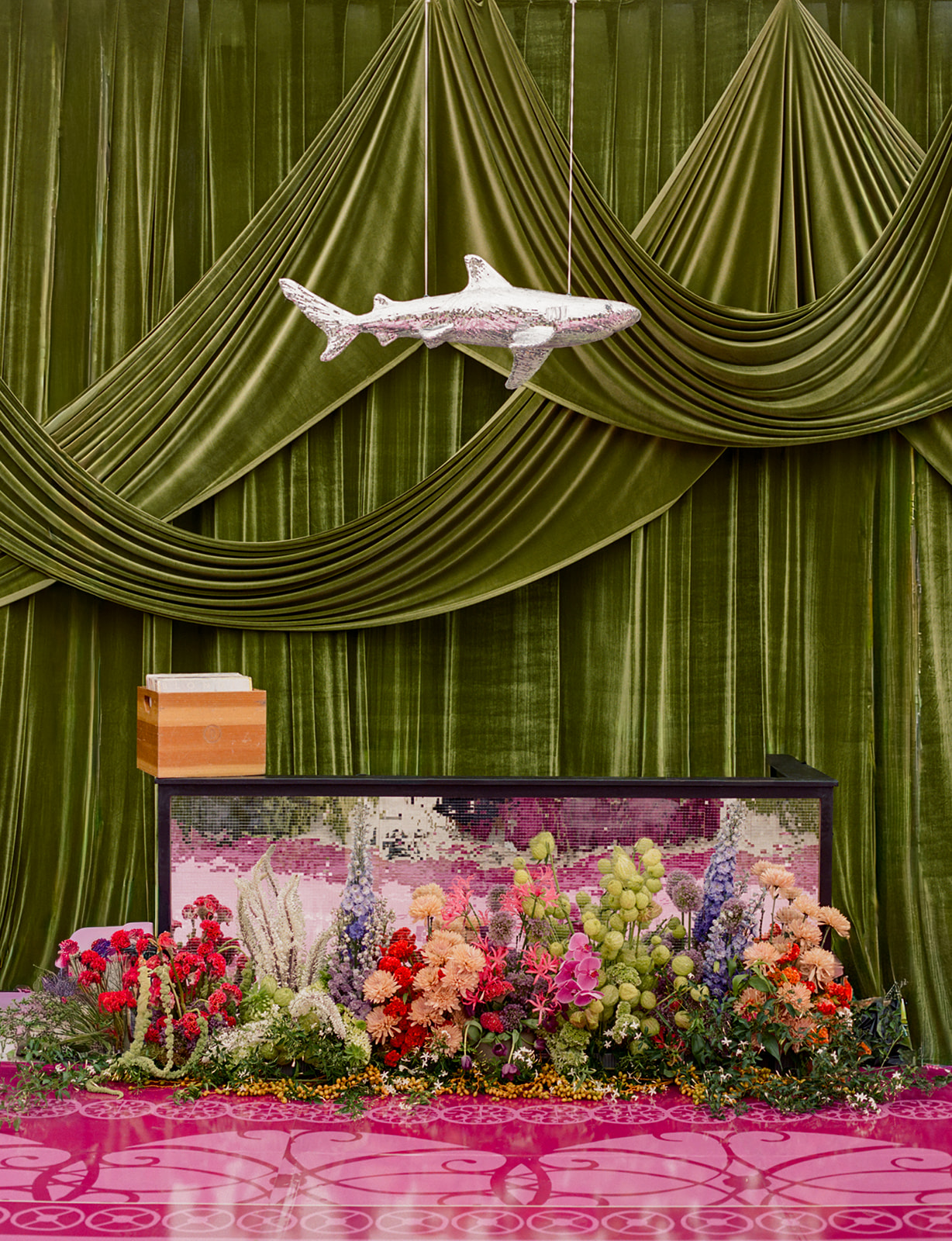 Decorative scene with green drapes, a white crocodile figure, a small stack of books, and a colorful floral arrangement on a pink surface.