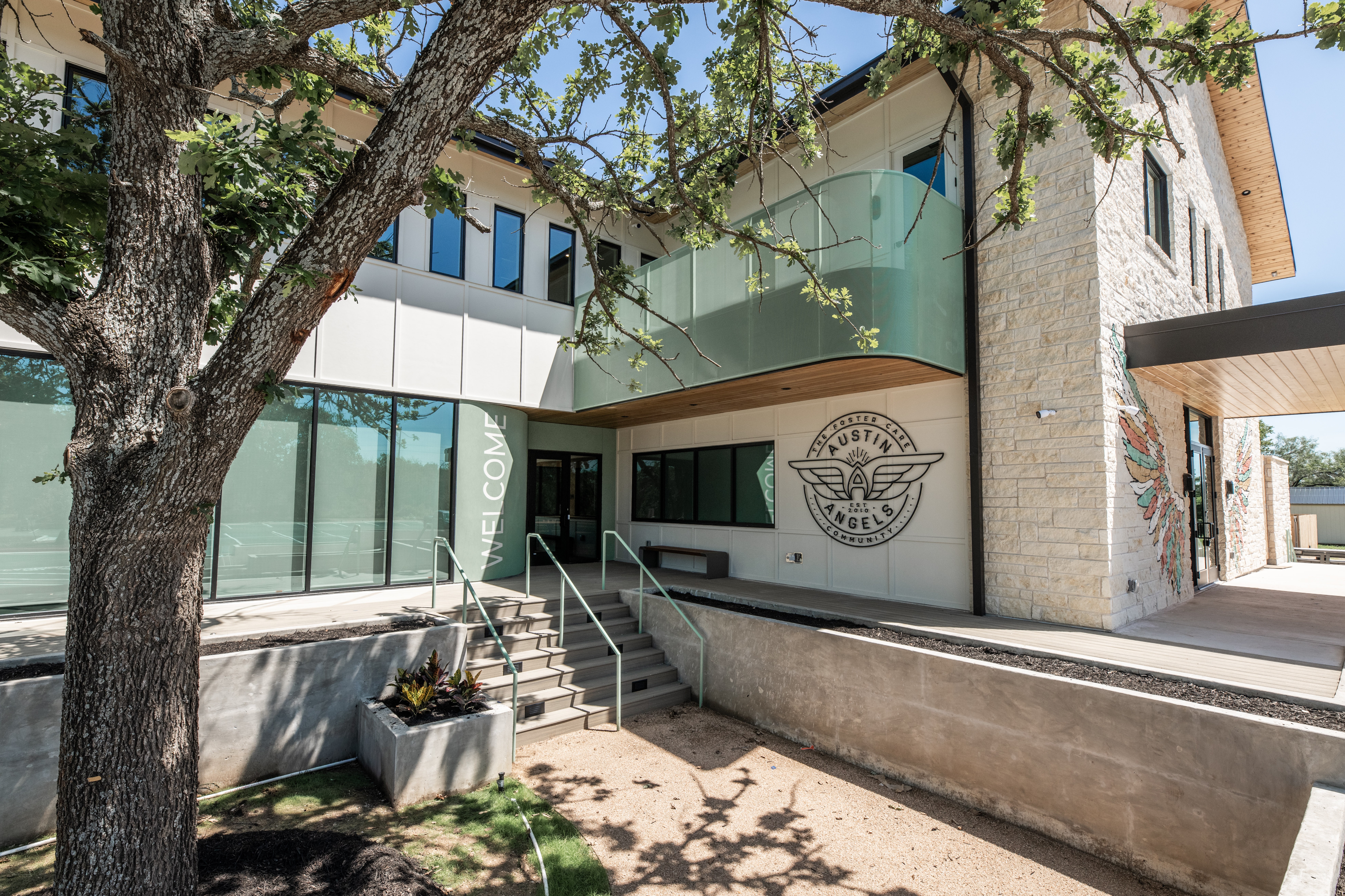 Building with glass windows, a tree in front, and a logo on the wall, with stairs and a ramp outside.
