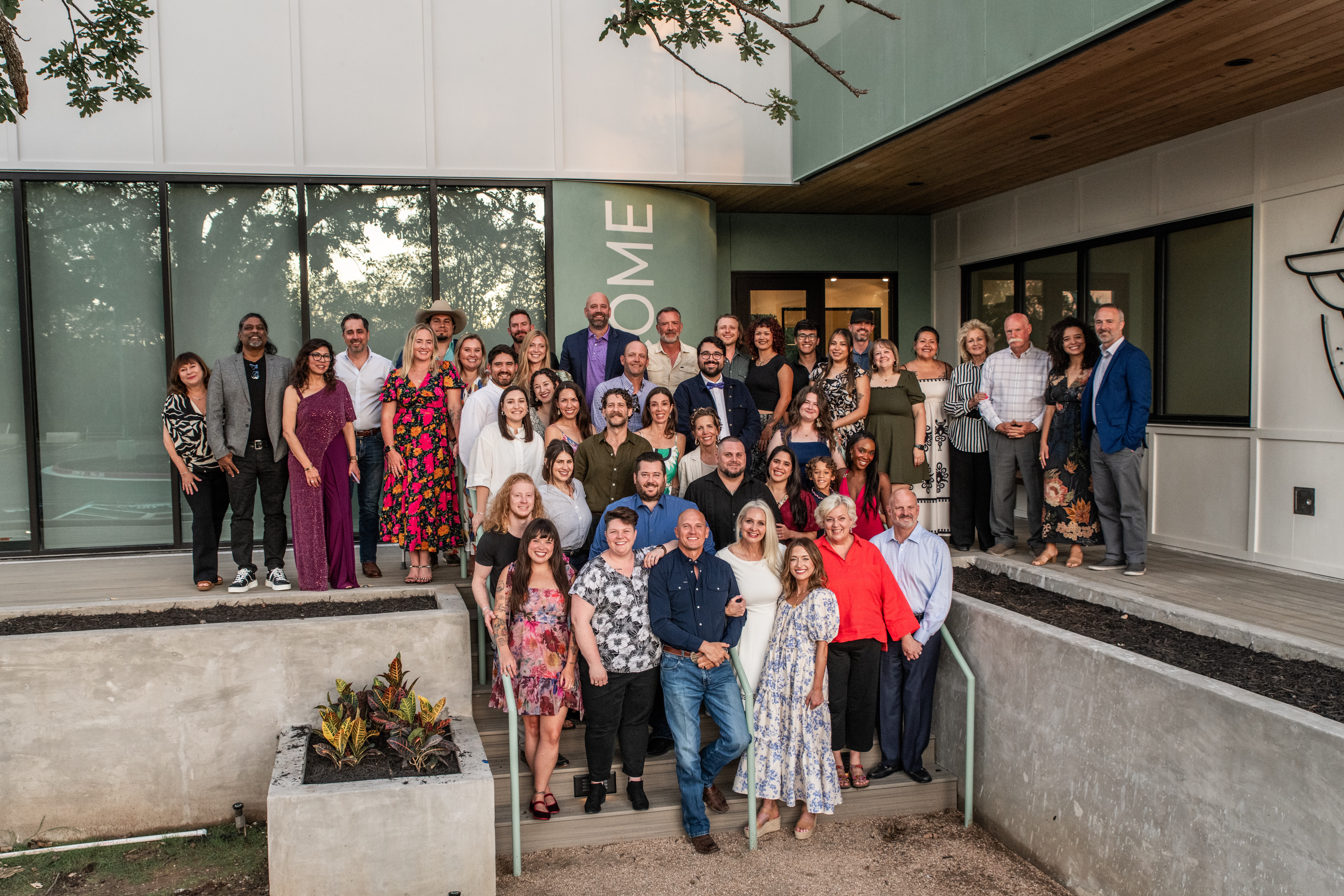 Group of people gathered outside a modern building, posing for a photo, with some holding balloons, in daytime.