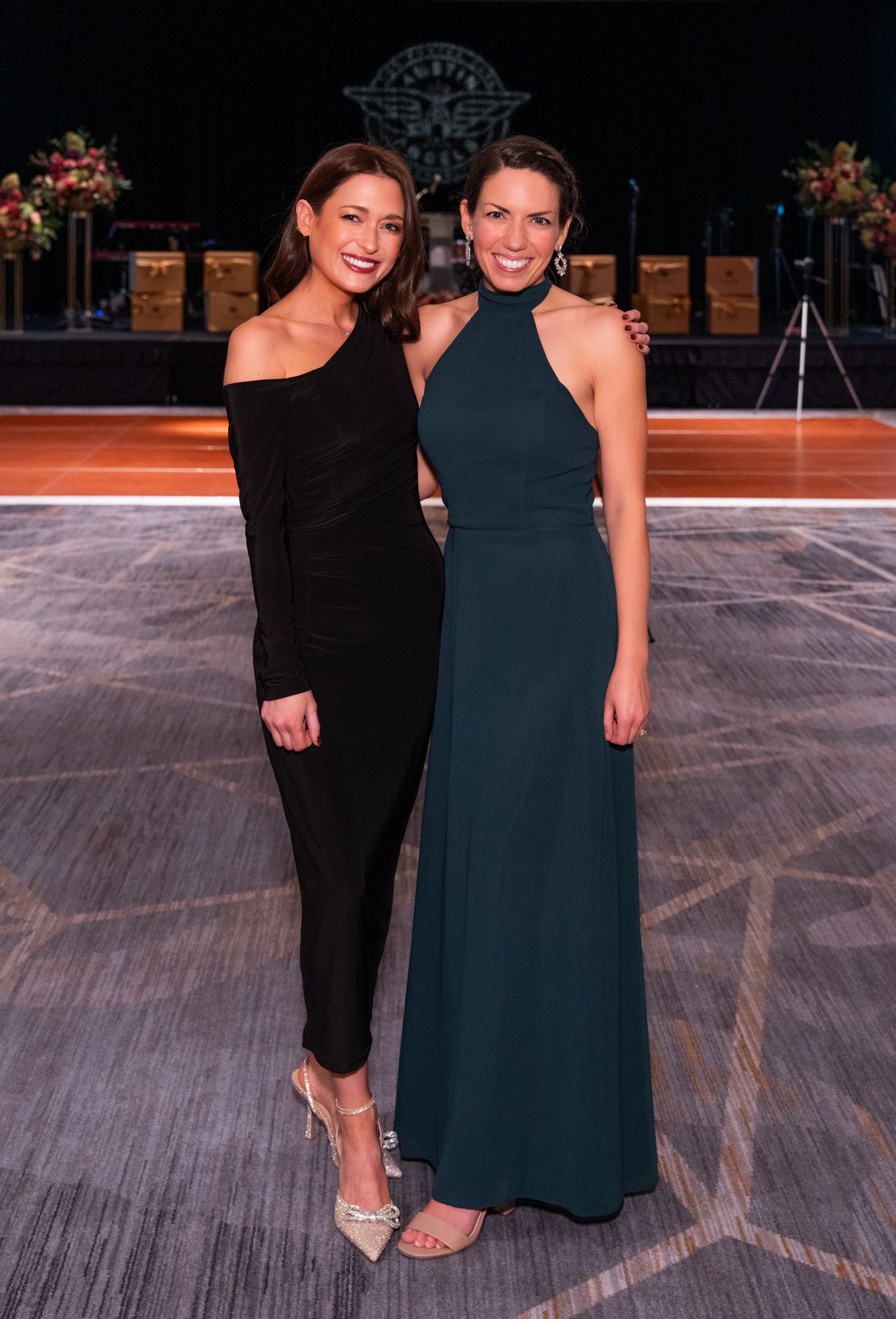 Two women in formal dresses standing on a stage with a dark background and floral arrangements.