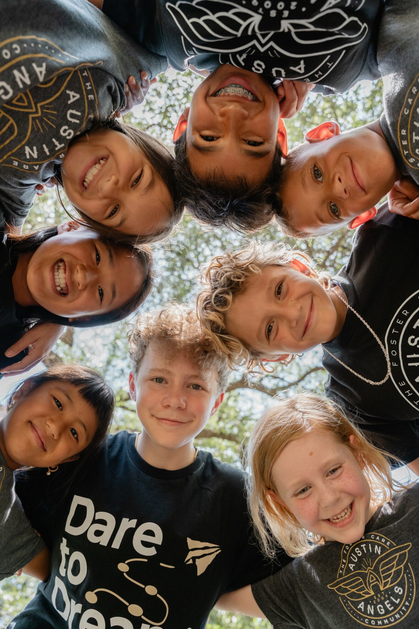 Group of children and a woman in a circle outdoors, smiling and looking down at the camera, trees in background.