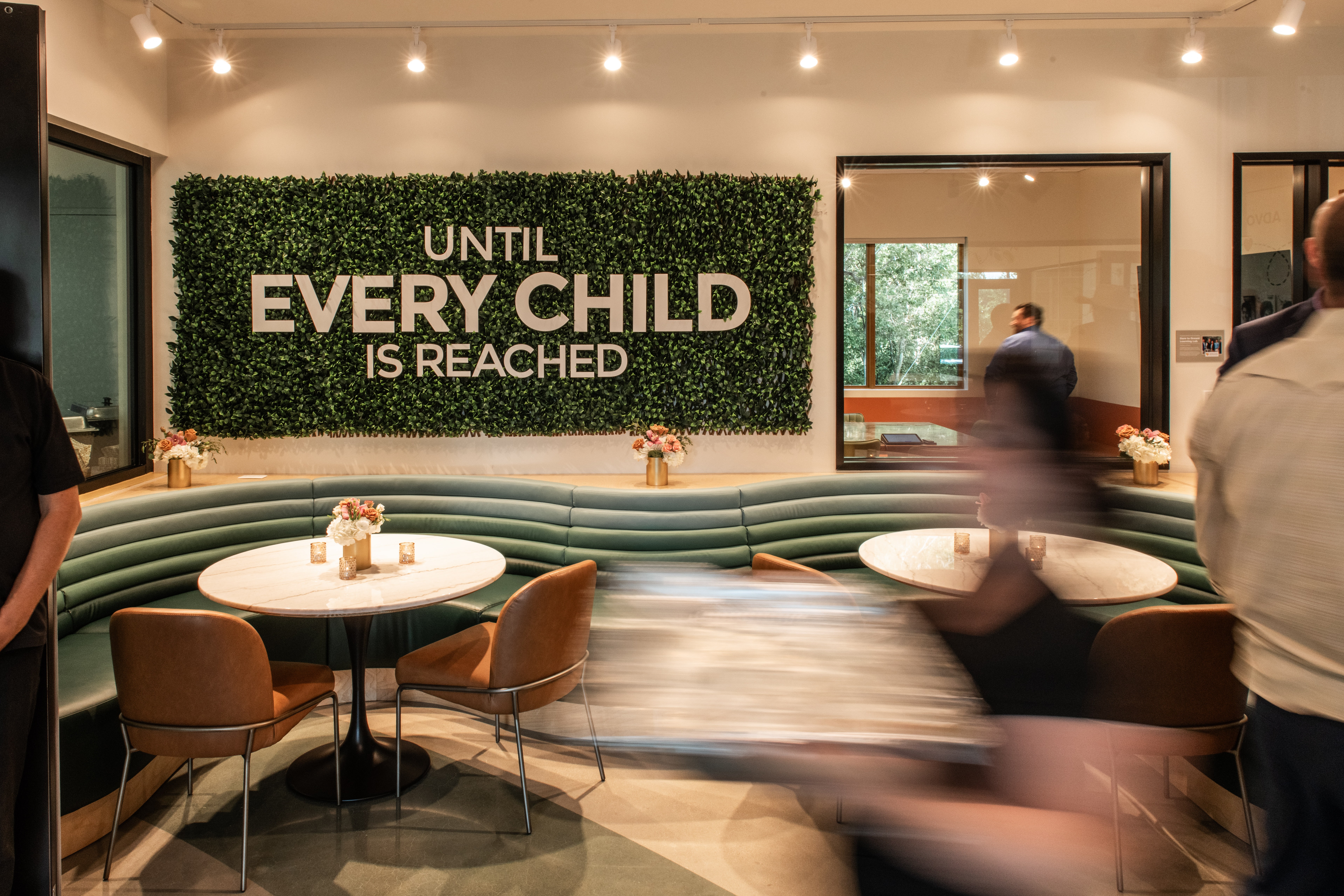 Interior of a cafe with a green wall sign reading 'Until Every Child is Reached,' tables, chairs, and people, some blurred motion.