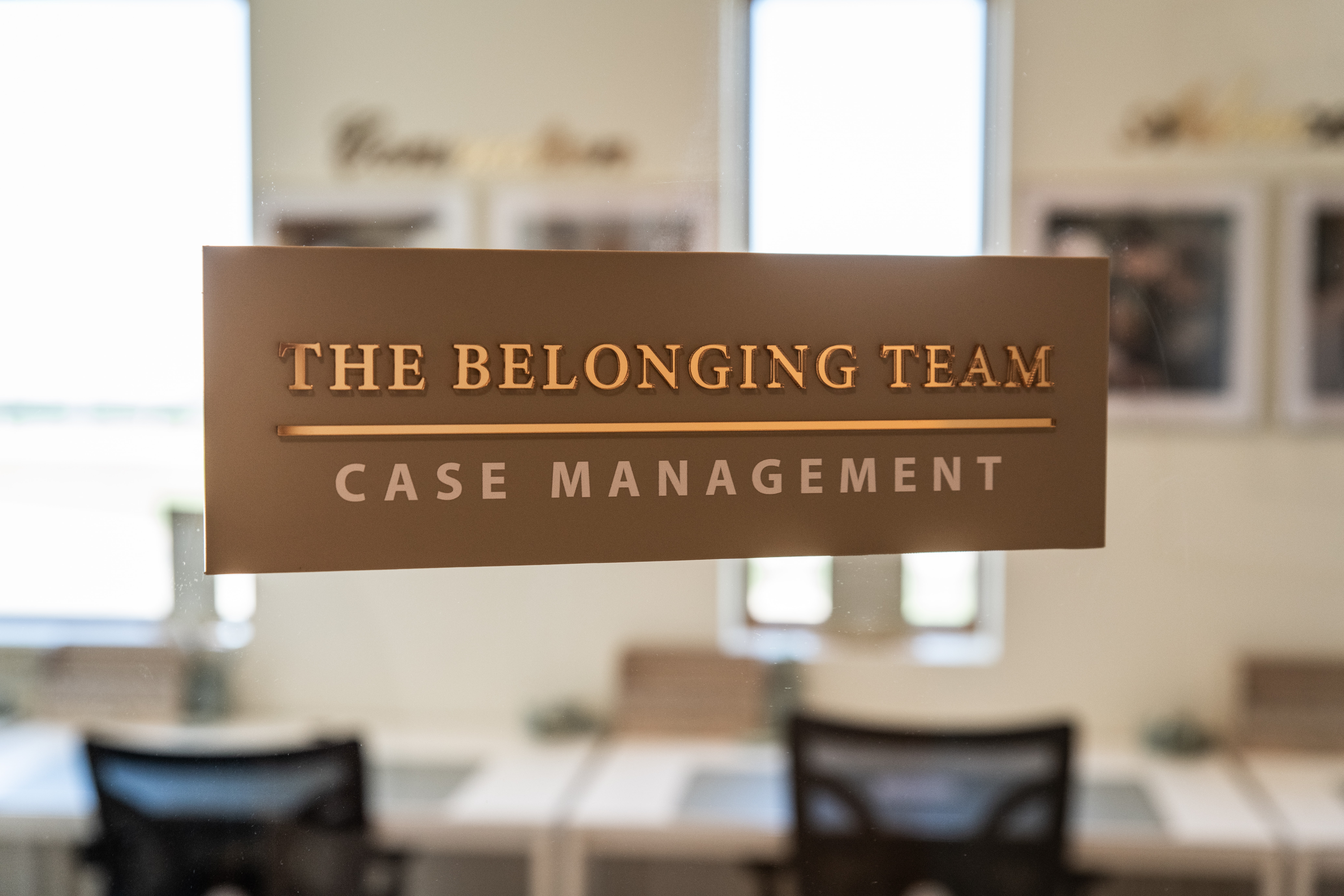 Sign reading 'The Belonging Team' and 'Case Management' in a room with tables and chairs, windows in background.