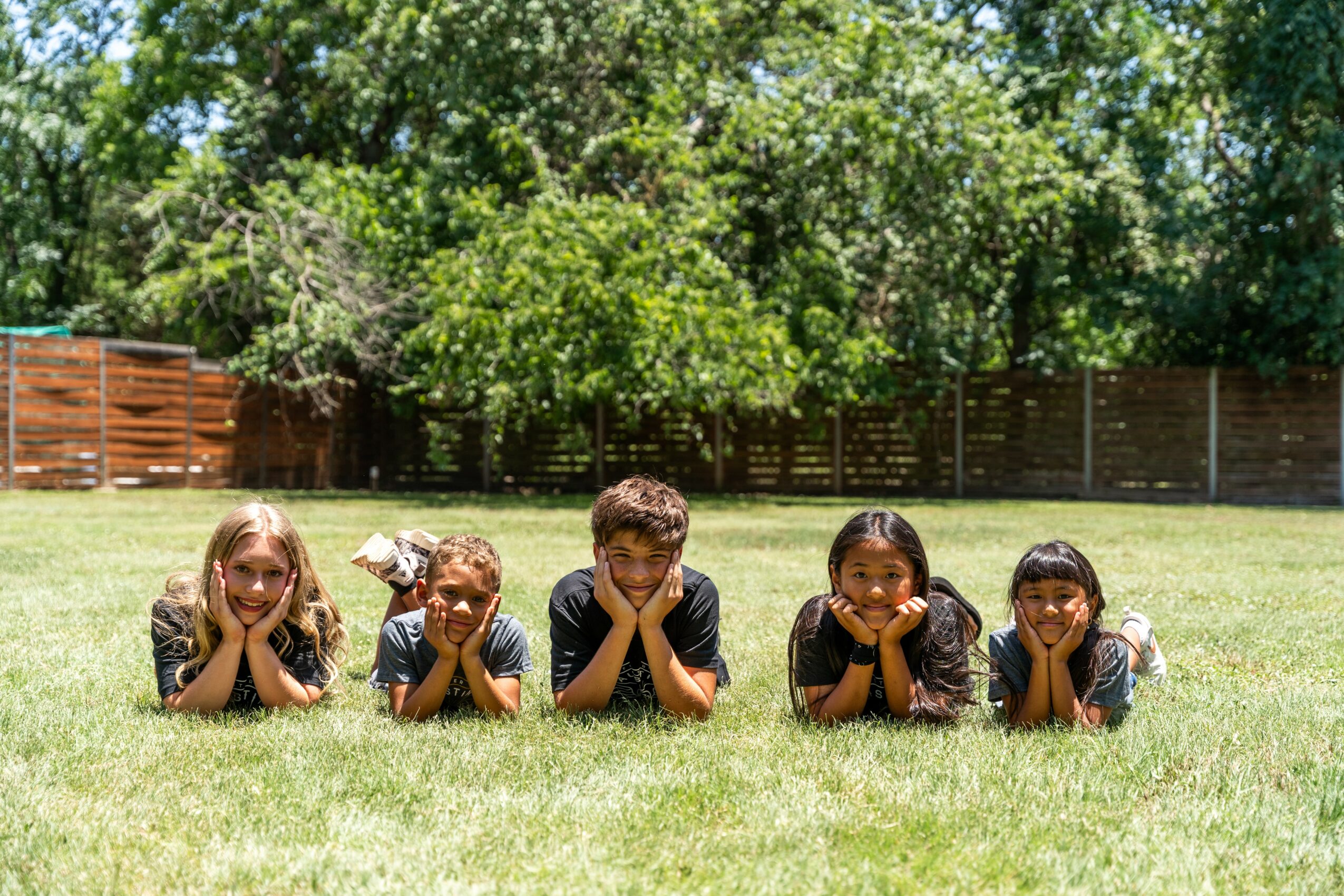 Five children lying on grass with heads resting on hands, outdoors with trees and a wooden fence in background.