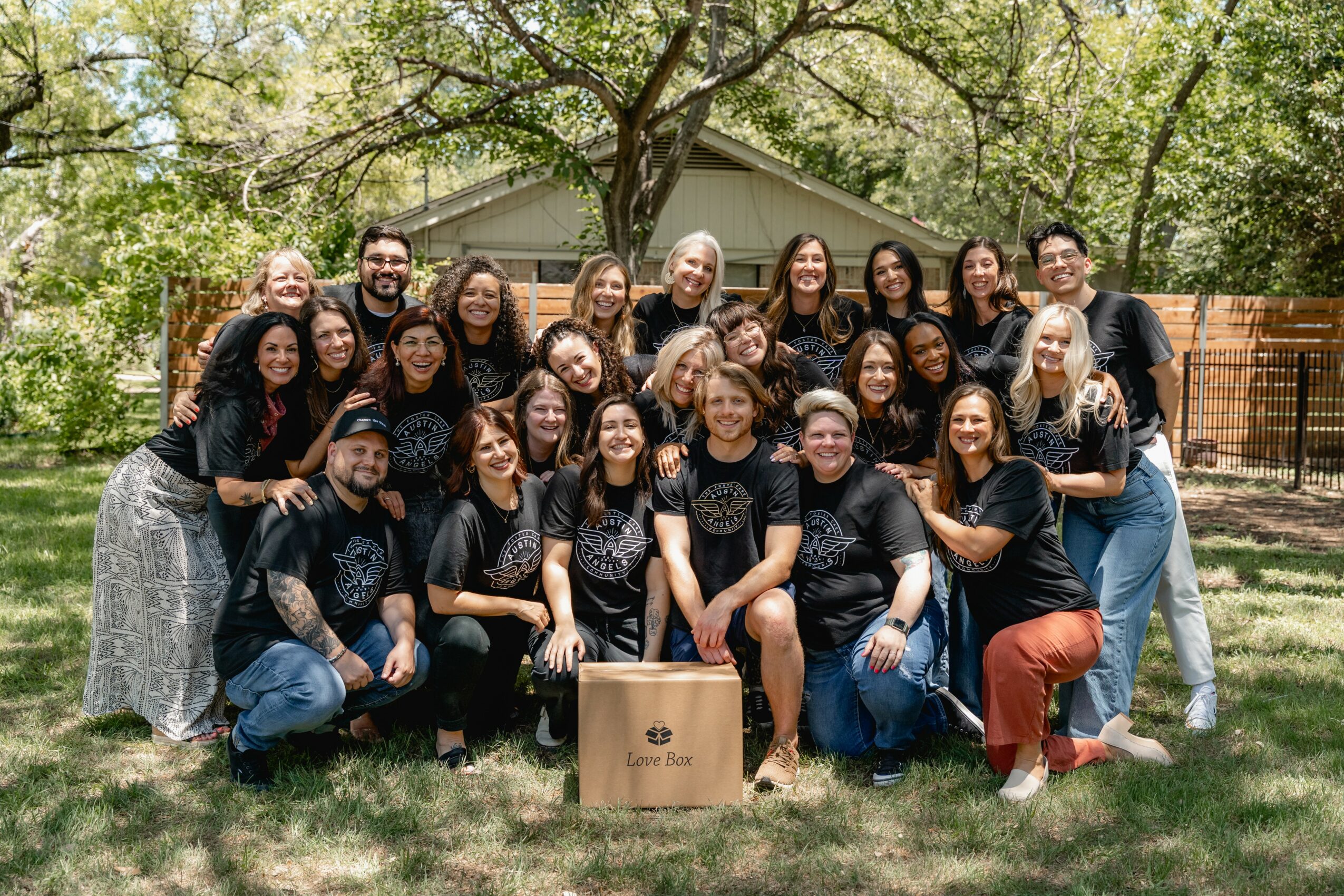 Group of 20 people outdoors, smiling, posing with a cardboard box, trees and a house in background.
