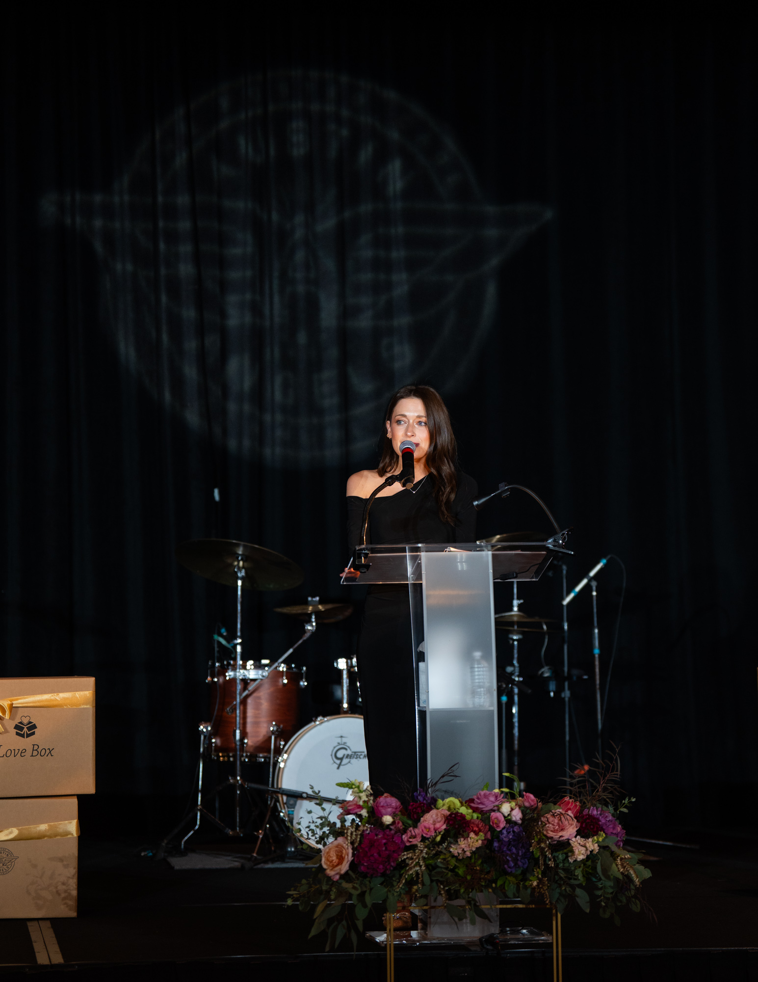 Woman speaking at a podium on stage with musical instruments and floral arrangement, dark background with circular projection, in a formal event.