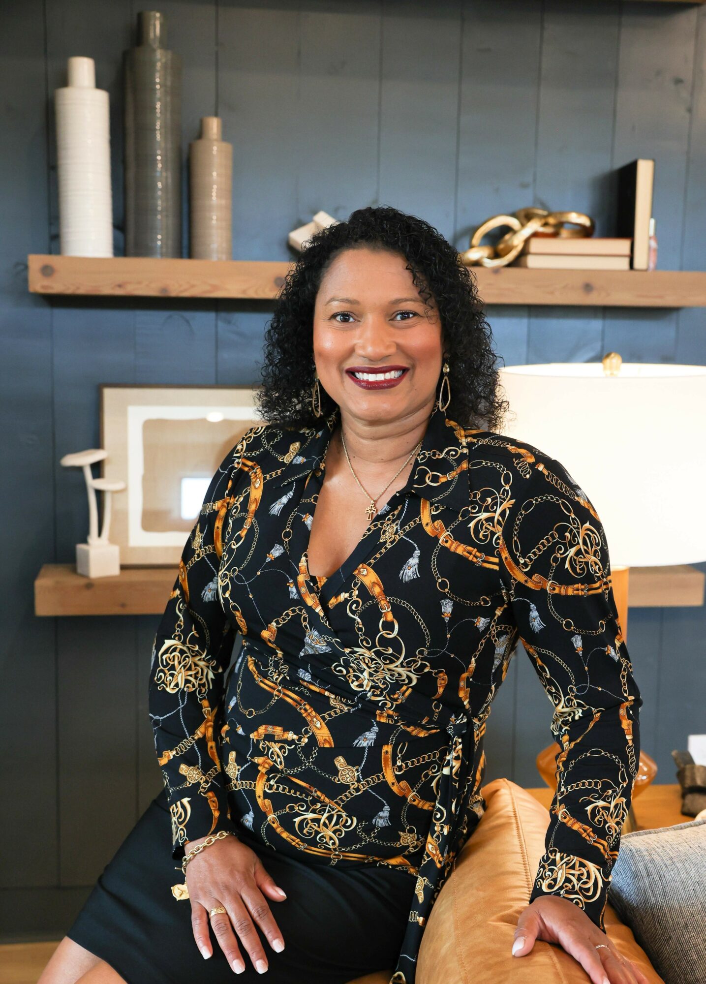 Smiling woman with curly hair wearing a patterned blouse, sitting in a room with decorative items on shelves behind her.