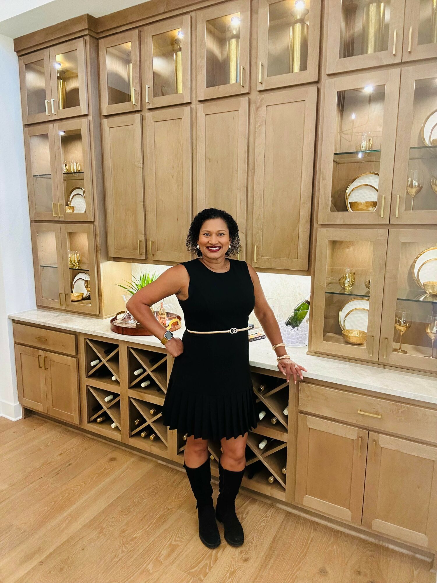 Woman in black dress and knee-high boots standing in front of wooden cabinet with glass display shelves, smiling.