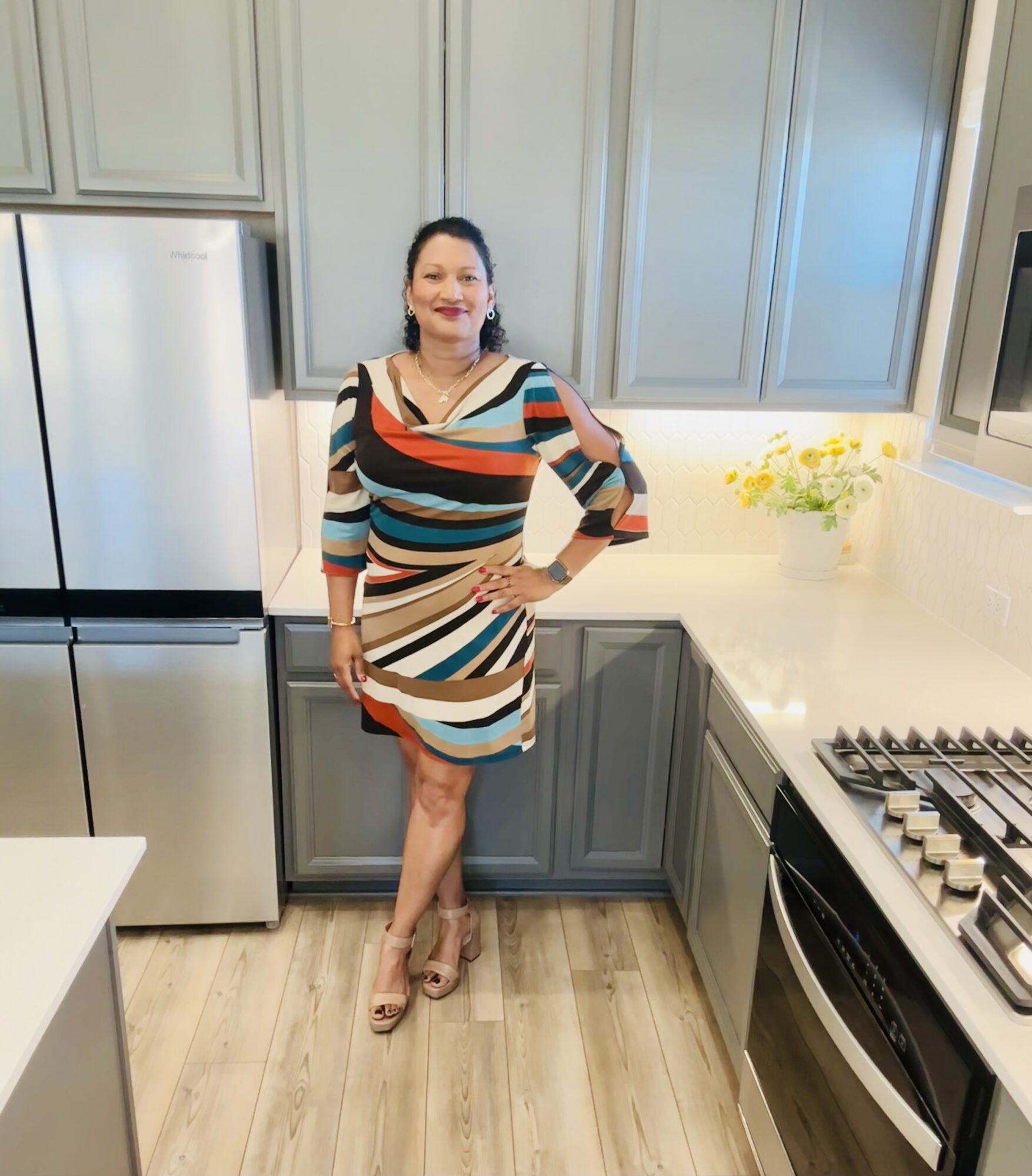 Woman standing in a kitchen with gray cabinets, white countertop, and a flower vase, smiling, wearing a colorful striped dress.