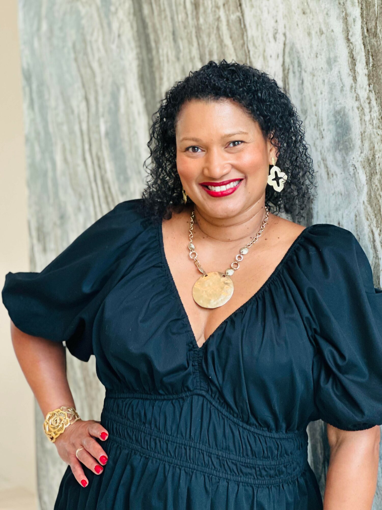 Woman with curly hair wearing a black dress, gold jewelry, and red lipstick, smiling in front of a marble background.