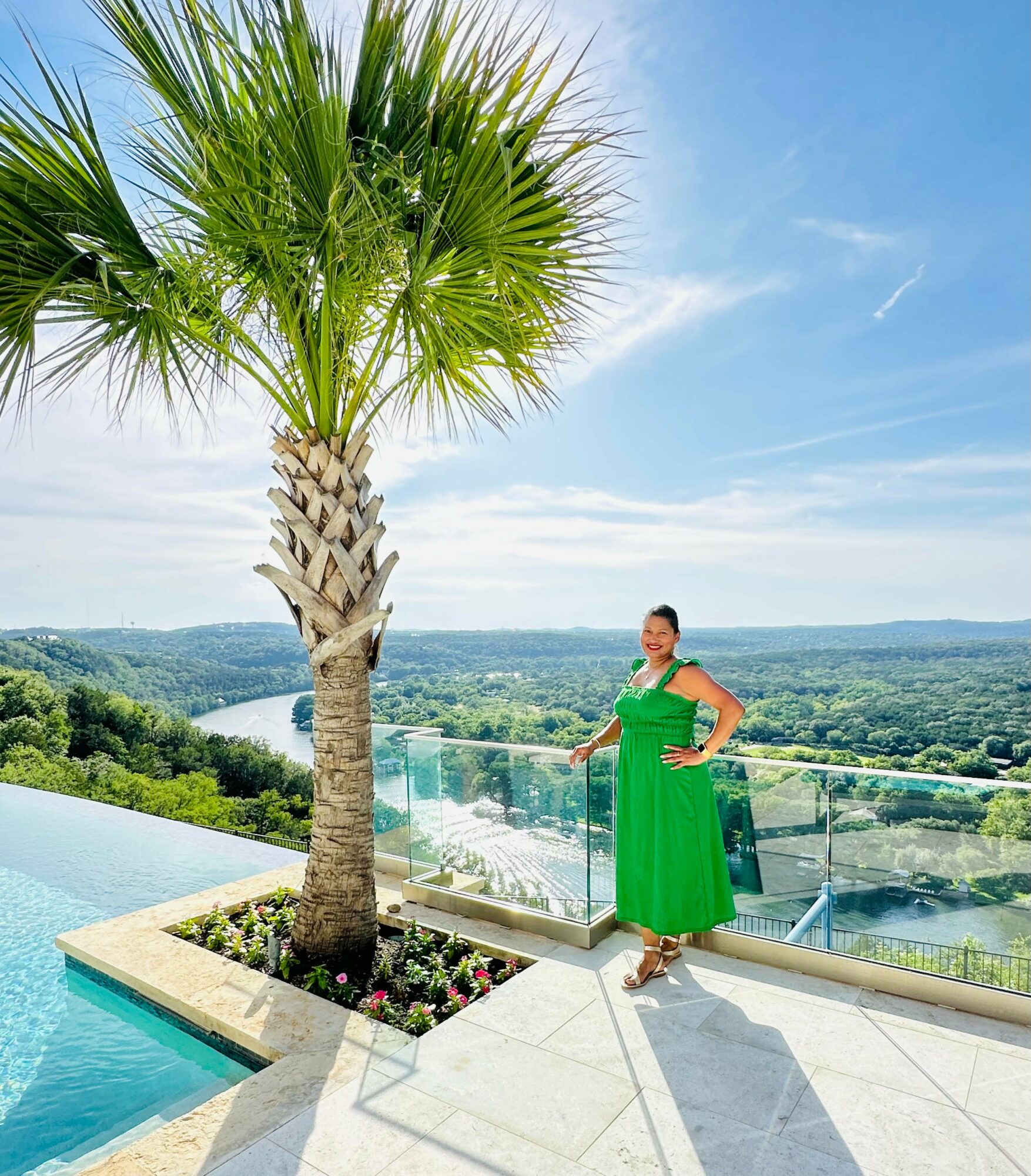 Woman in green dress standing on balcony with a large palm tree and river view behind her.