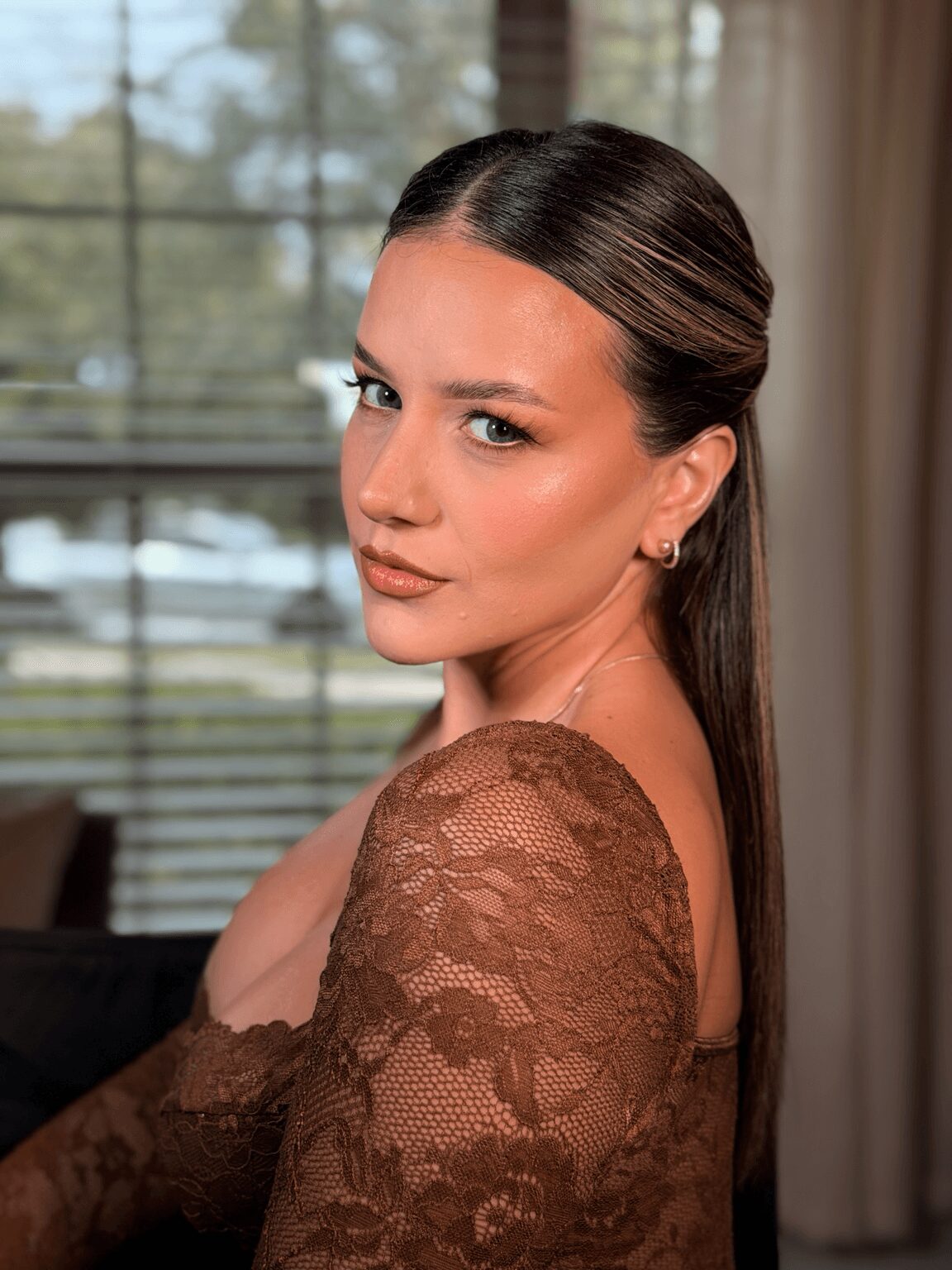 Young woman with long dark hair and makeup, wearing a brown lace top, standing indoors near a window with blinds.