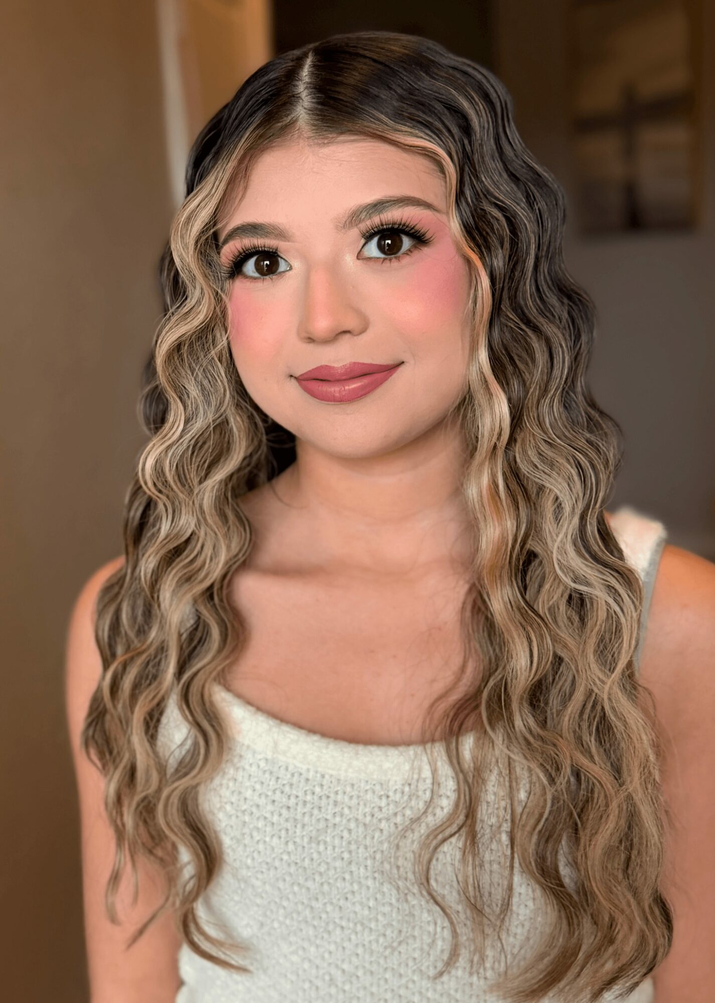 Young woman with long, wavy hair and makeup, wearing a white top, smiling softly indoors.