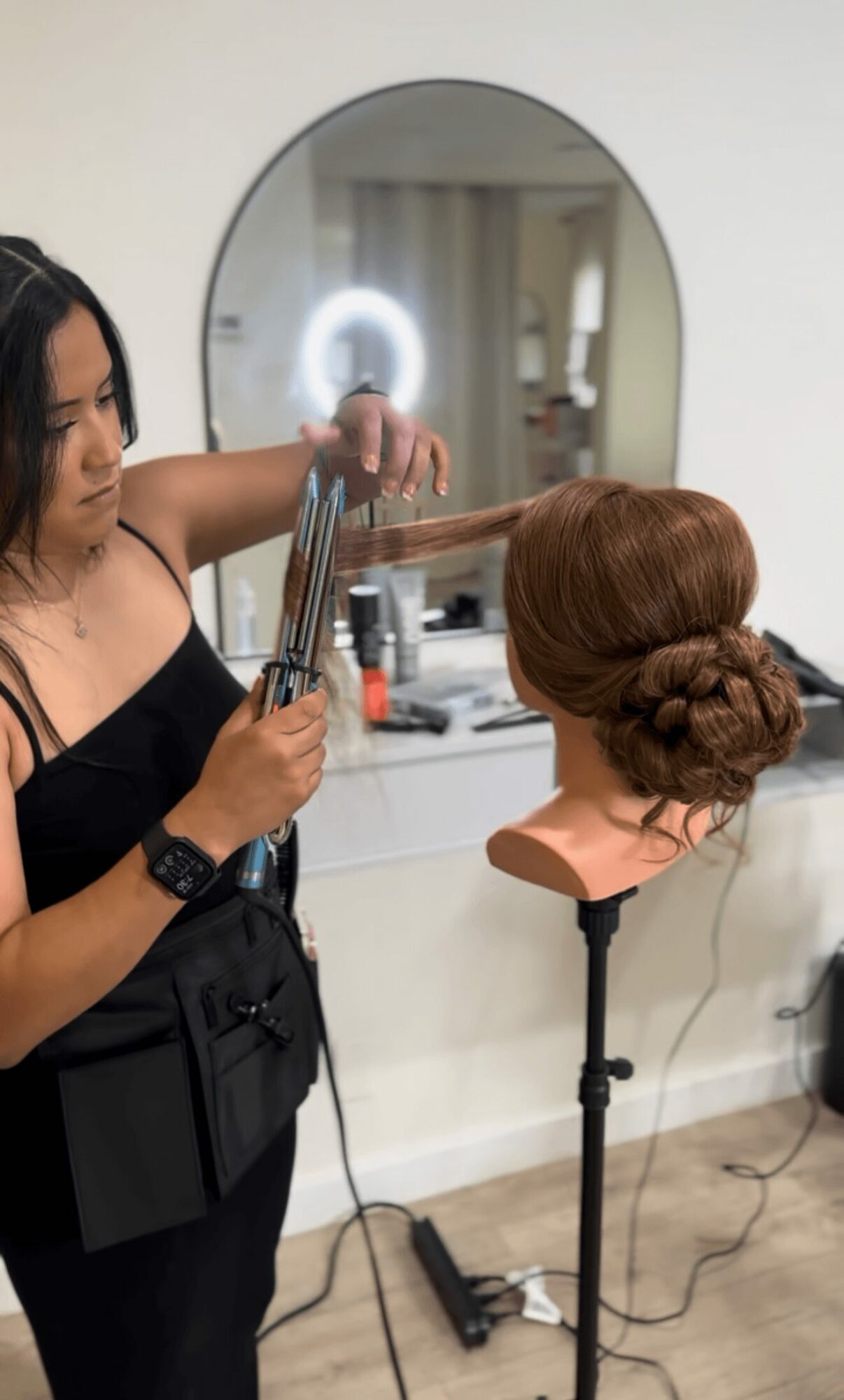 A hairstylist working on a mannequin head with styled hair, in front of a mirror in a salon, with tools nearby.