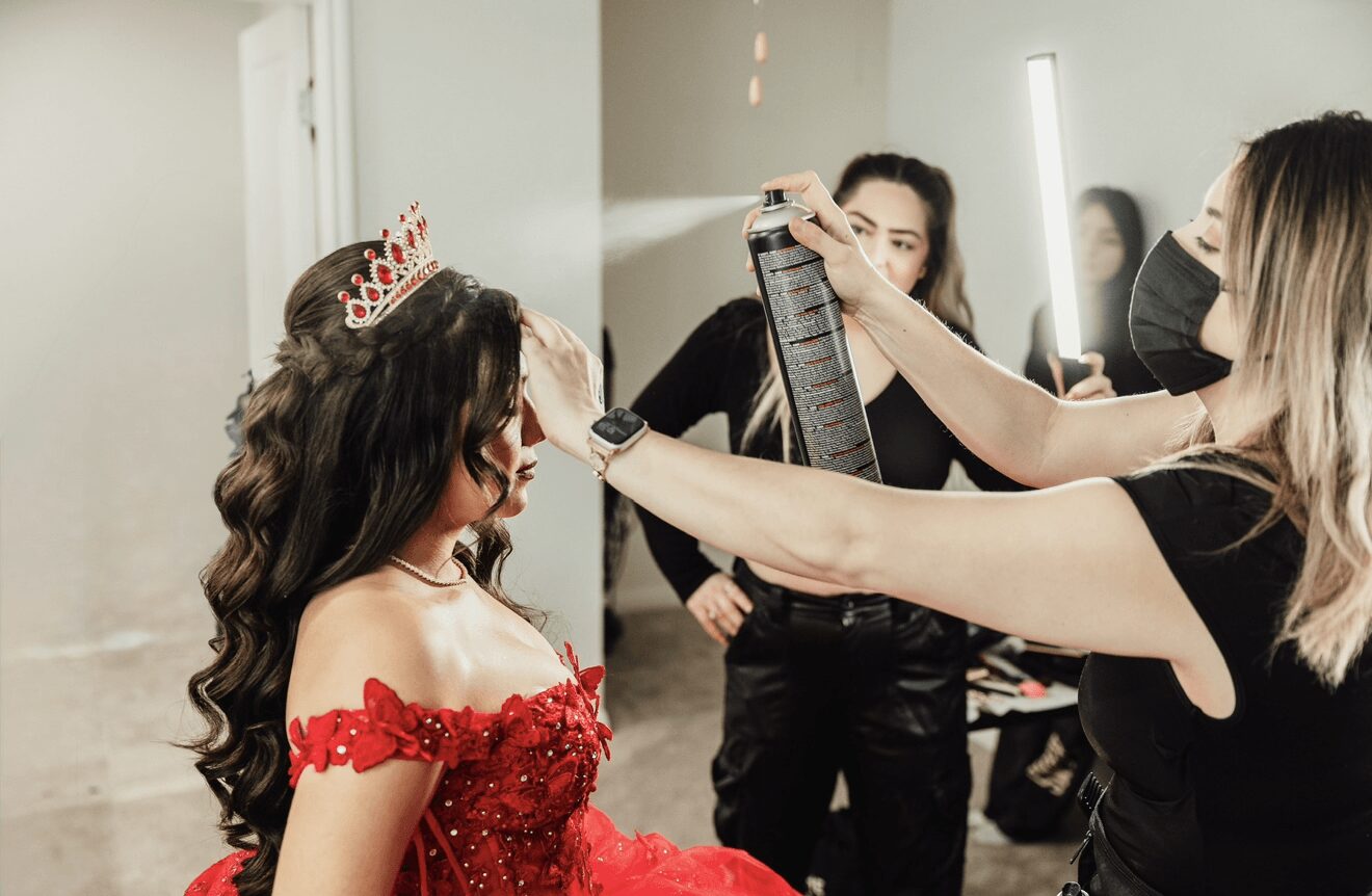 A woman in a red dress and tiara is being styled by a makeup artist in a mirror, with others observing.