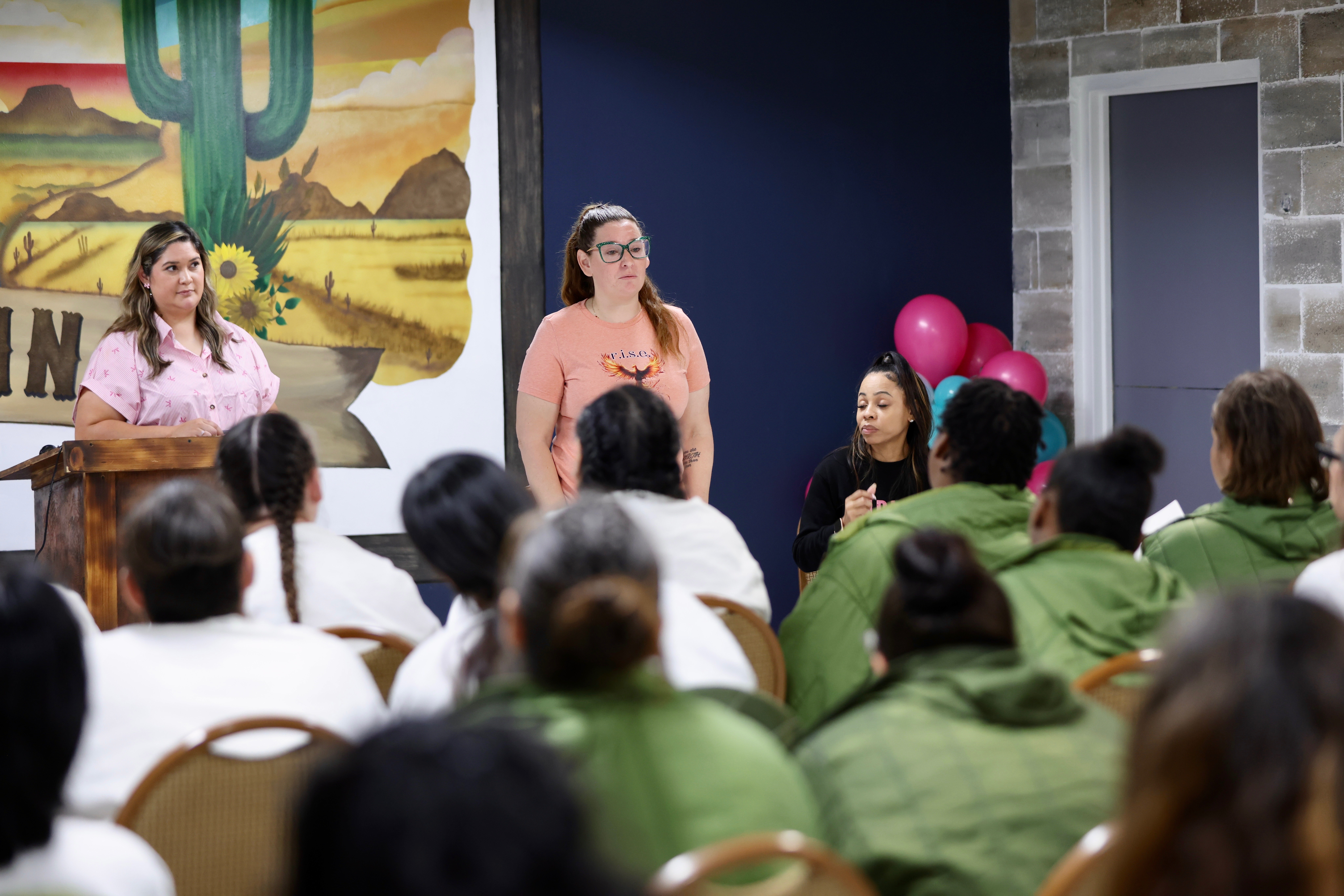 Three women speak to an audience in a room with a desert mural and pink balloons, audience members seated and listening.