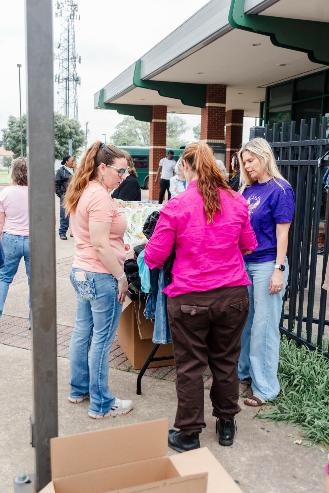 Group of four women gathered around a table outside a building, talking and looking at items, with other people in background.