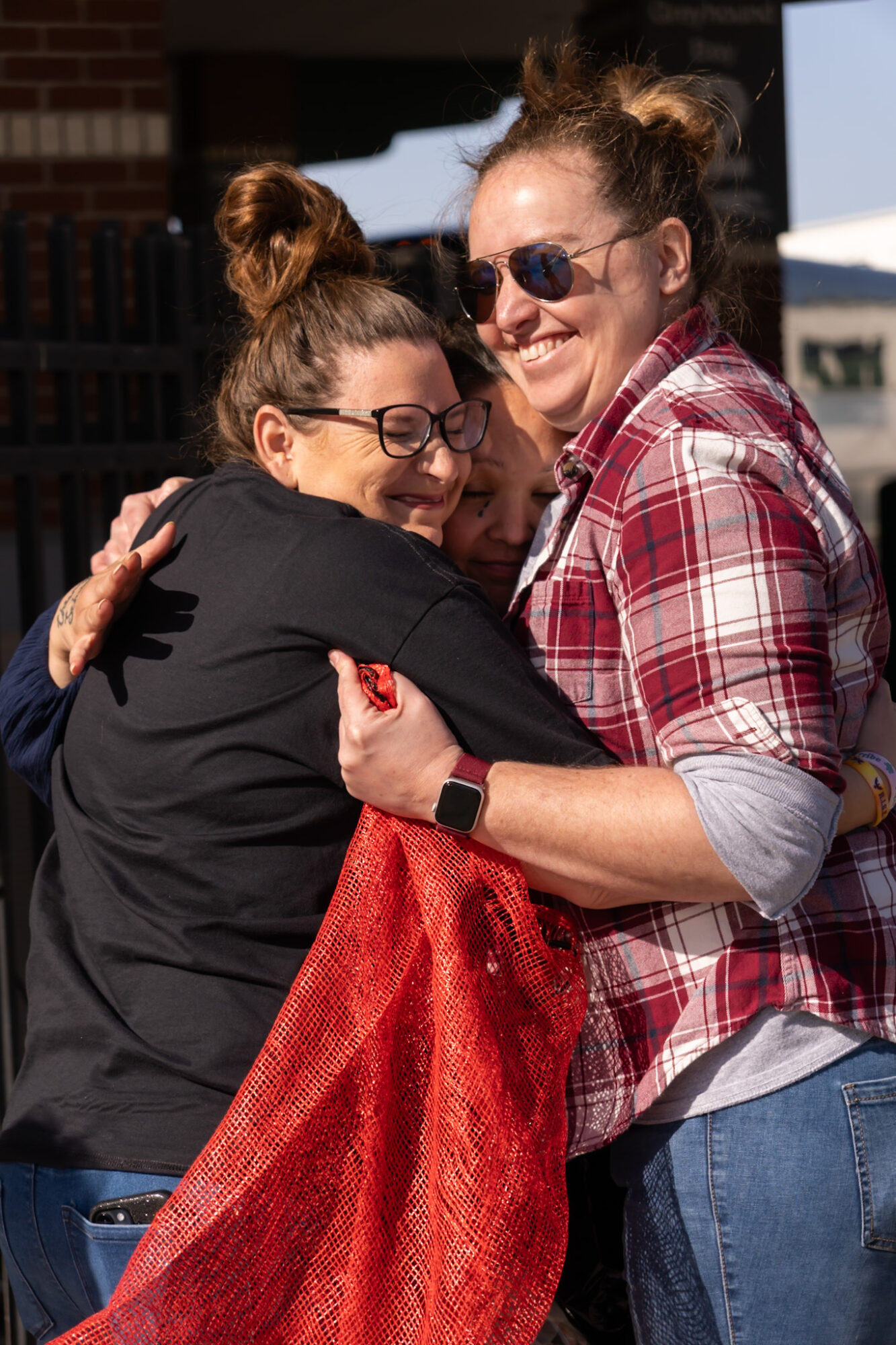 Two women hugging outdoors, smiling, one wearing sunglasses and a plaid shirt, the other with glasses and a black jacket.