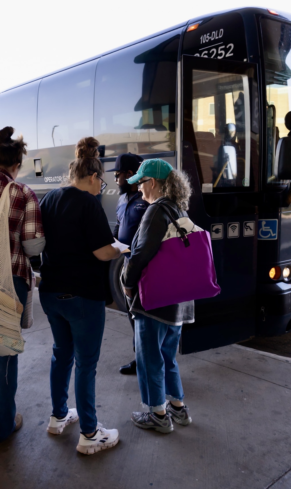 Group of people standing near a bus, some wearing backpacks and hats, on a concrete surface.